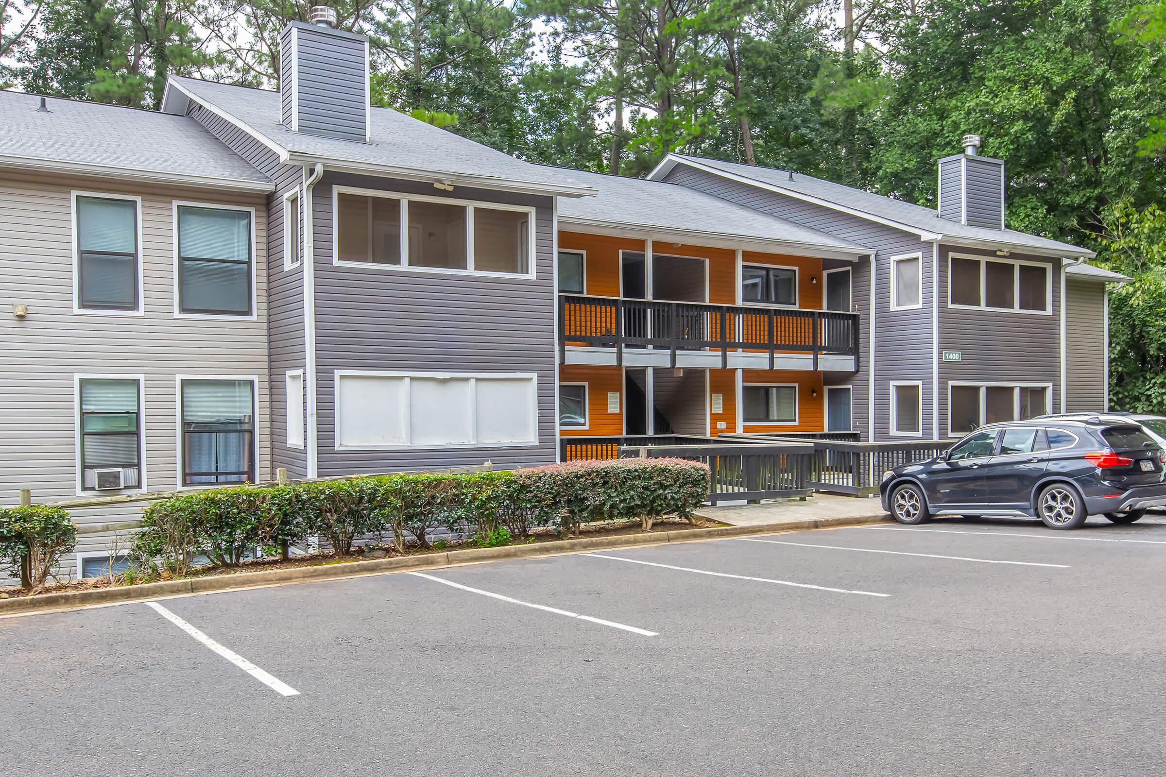 A multi-unit apartment building with a mix of gray and orange accents. The structure has two levels with balconies and several windows. A parking lot is visible in front, with one parked vehicle and landscaped bushes along the driveway. Trees are in the background, contributing to a natural setting.