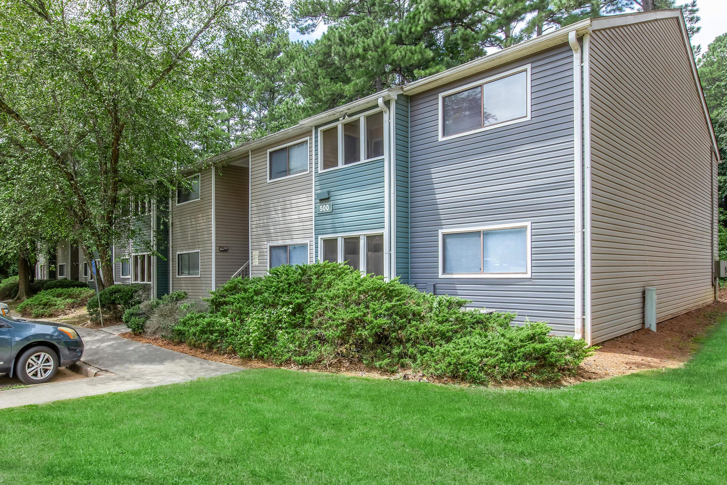 A two-story apartment building with blue and grey siding, surrounded by green grass and bushes. A parked car is in the foreground, and tall trees are visible in the background, indicating a residential area. The building features multiple windows and a simple architectural design.