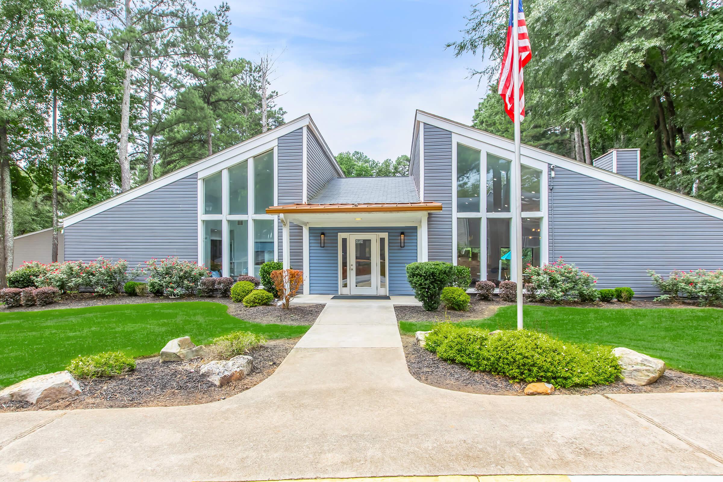 Modern building with a unique architectural design featuring large glass windows and a sloped roof. The entrance is framed by neatly landscaped greenery and flowers. An American flag stands prominently at the front, adding a touch of patriotism to the scene.