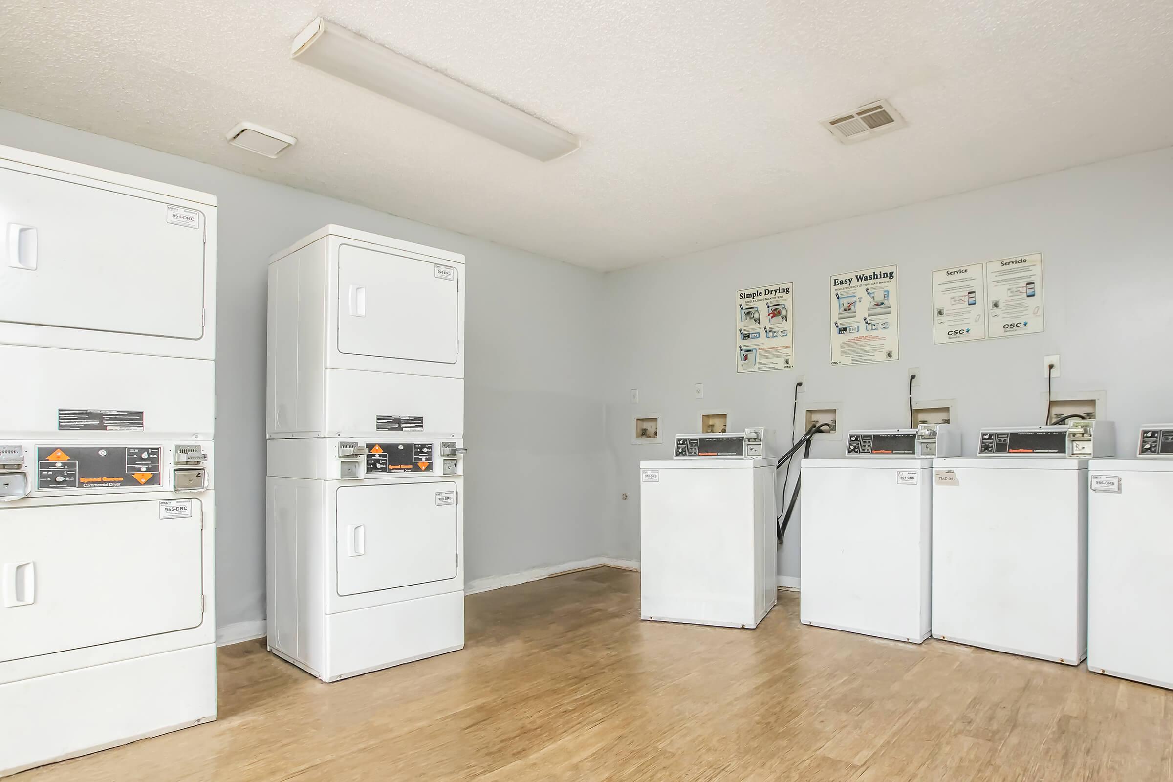 A clean laundry room featuring several stacked and front-loading washing machines and dryers. The space is well-lit with light-colored walls and wooden flooring. There are also instructional signs on the wall regarding laundry use and settings.