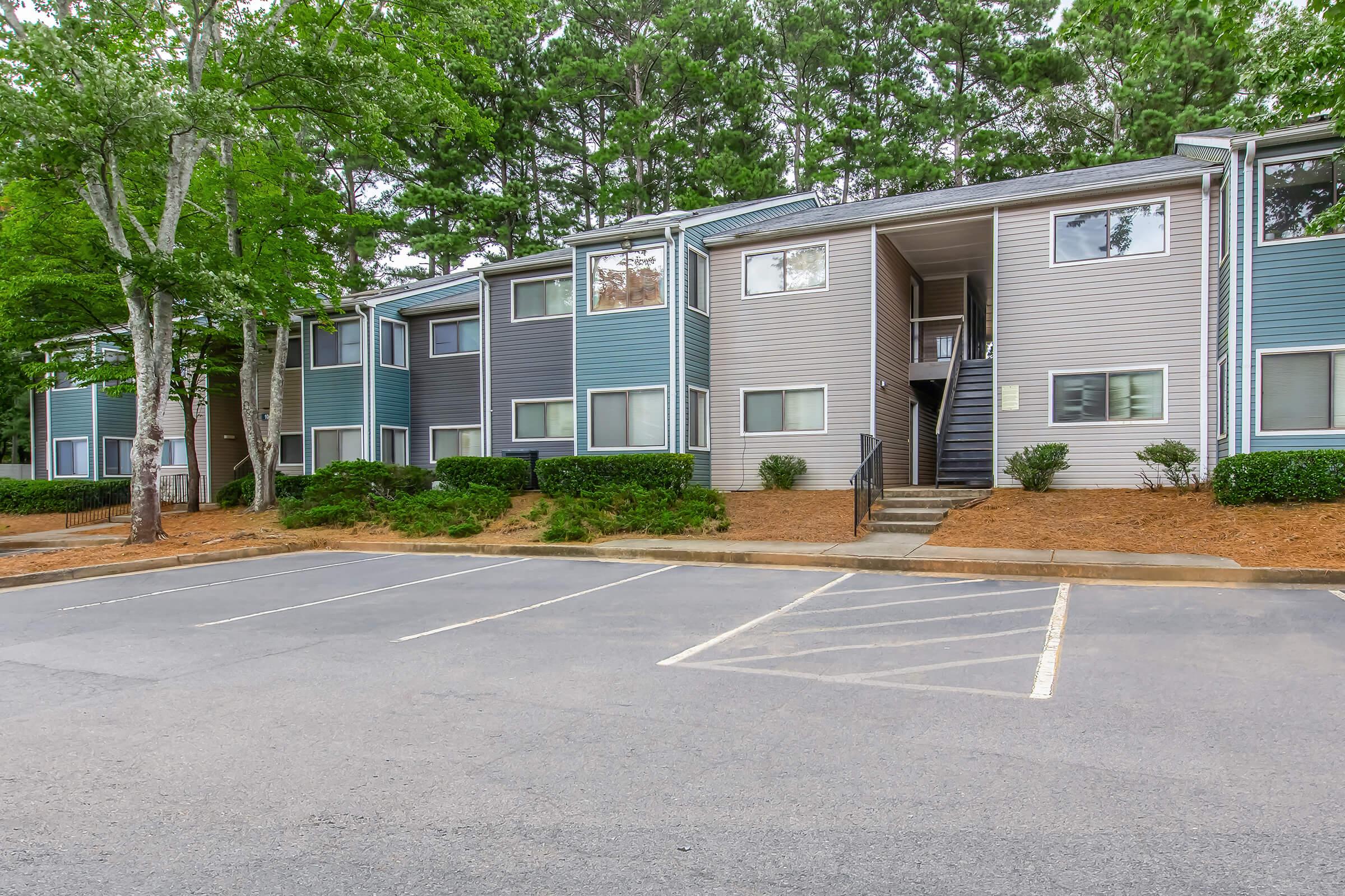 A view of a multi-unit apartment building surrounded by trees. The exterior features a combination of colors, with several windows visible. A paved parking area is in front, with designated parking spaces, and landscaping including shrubs and wood mulch.