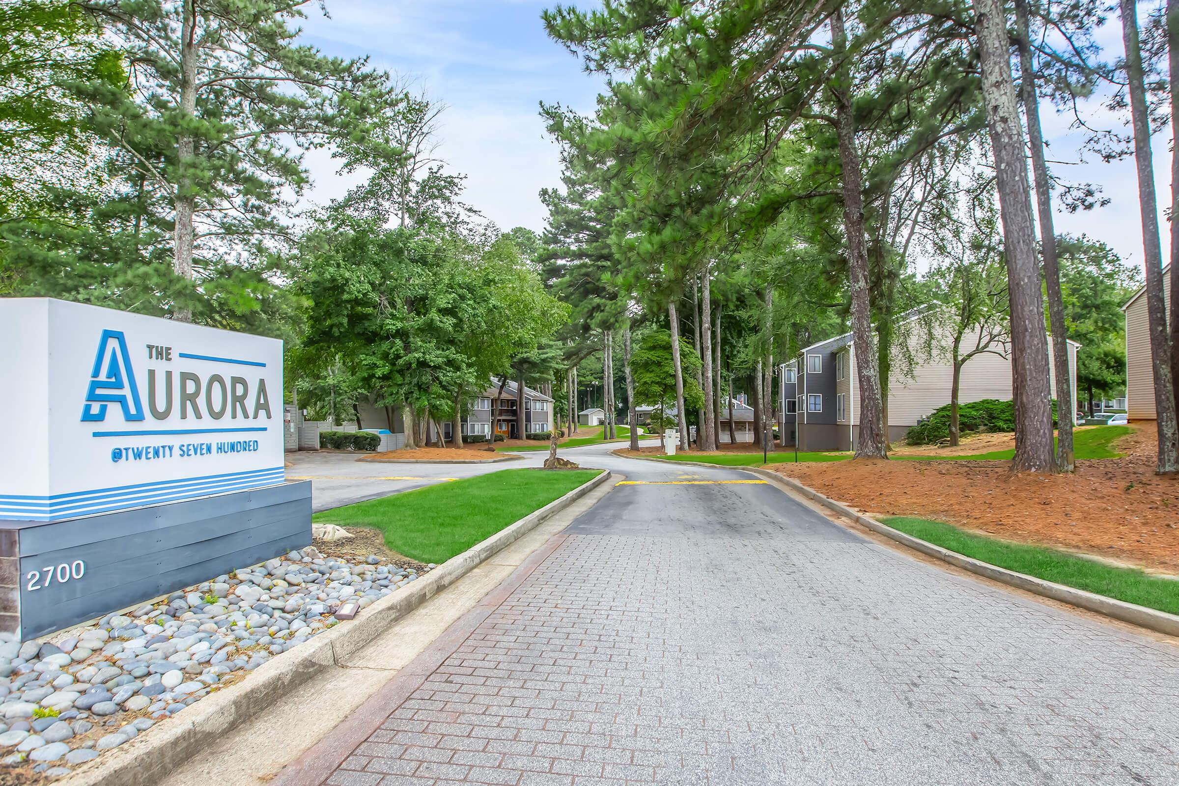 Sign for "The Aurora" at 2700, with a view of a curved driveway leading through a landscaped area featuring trees and shrubbery. Residential buildings are visible in the background, set among greenery. The path is clear and well-maintained, suggesting a welcoming community atmosphere.
