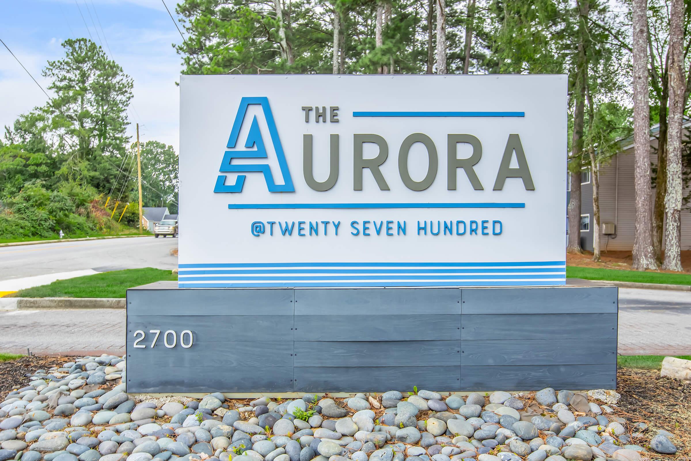 Sign for "The Aurora @Twenty Seven Hundred" located at 2700, featuring a modern design with blue and gray colors and surrounded by pebbles and greenery.