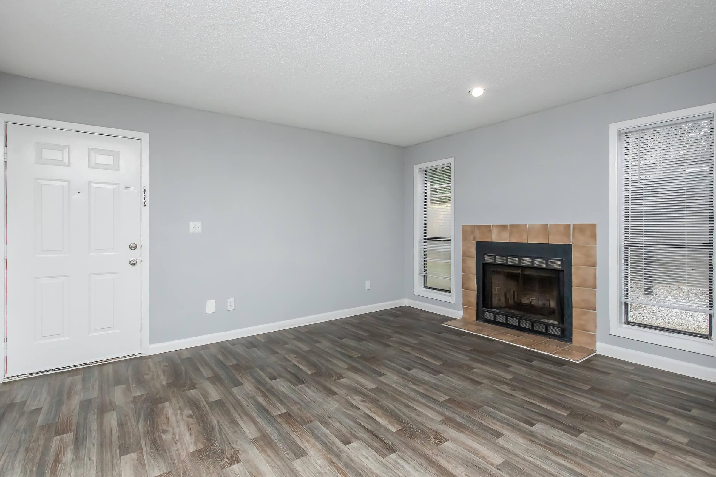 A spacious living room with light gray walls and wooden flooring. The room features a fireplace with a tiled surround and large windows allowing natural light. A front door is visible on the left, and the overall decor is modern and minimalist.