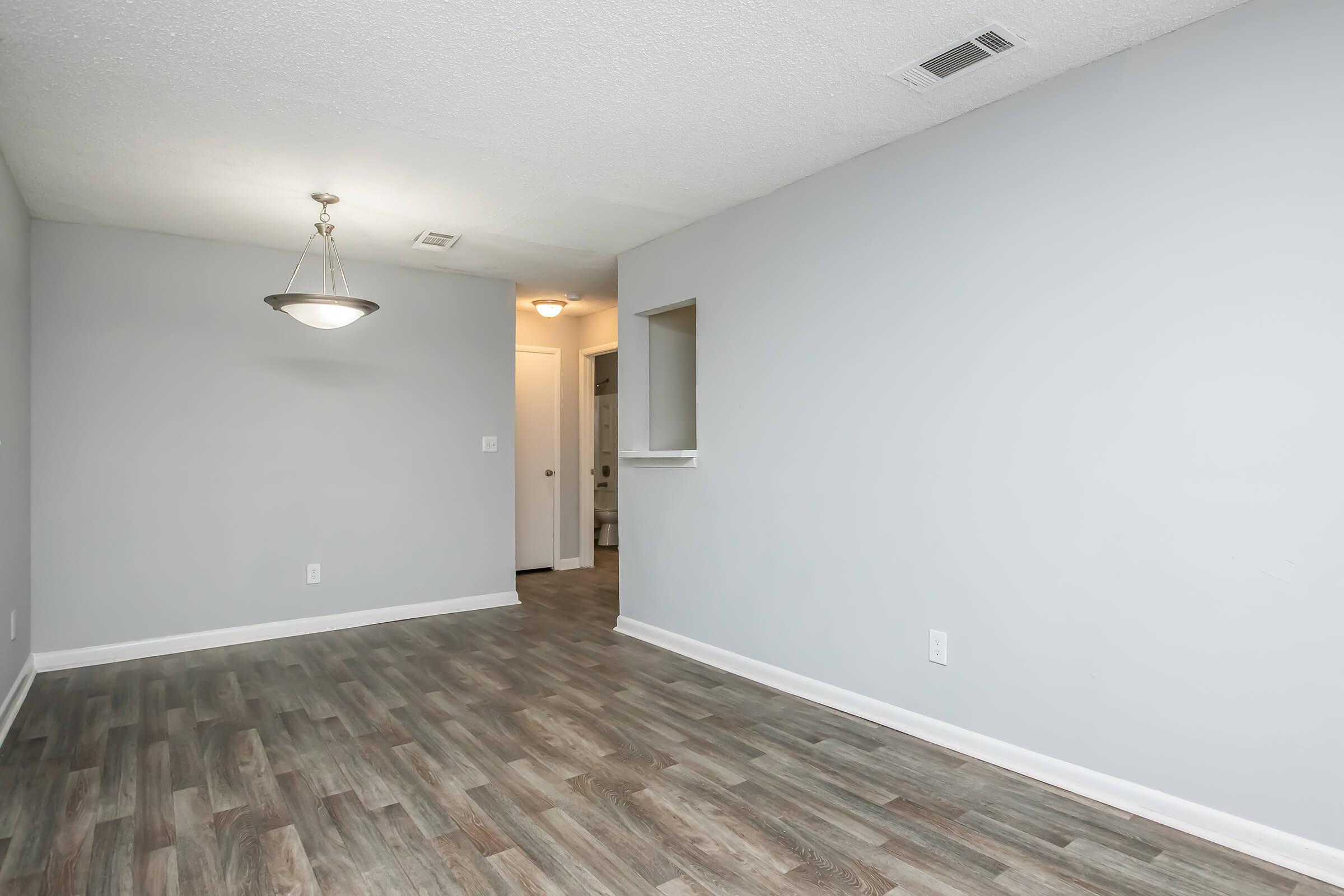 A spacious, empty room featuring light gray walls, a wood-like floor, and a ceiling light fixture. There's a small opening to another room on the right, and a doorway leading to a bathroom or hallway in the background. The space is well-lit and has a clean, modern appearance.