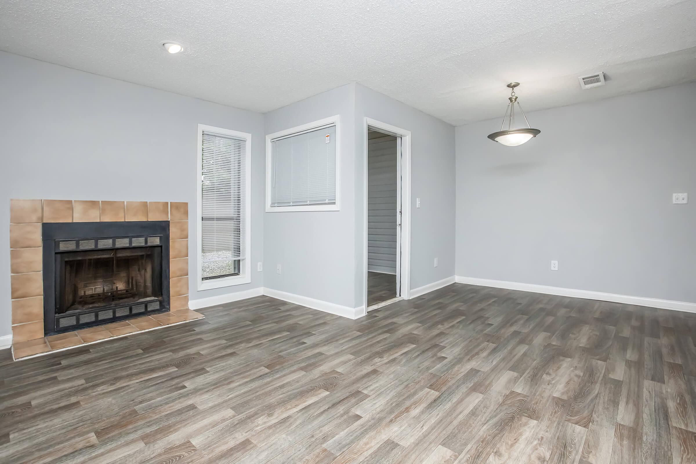 A spacious living area featuring light gray walls, a tiled fireplace, large windows with blinds, and modern laminate flooring. A doorway leads to another room, and a ceiling light fixture adds brightness to the space.