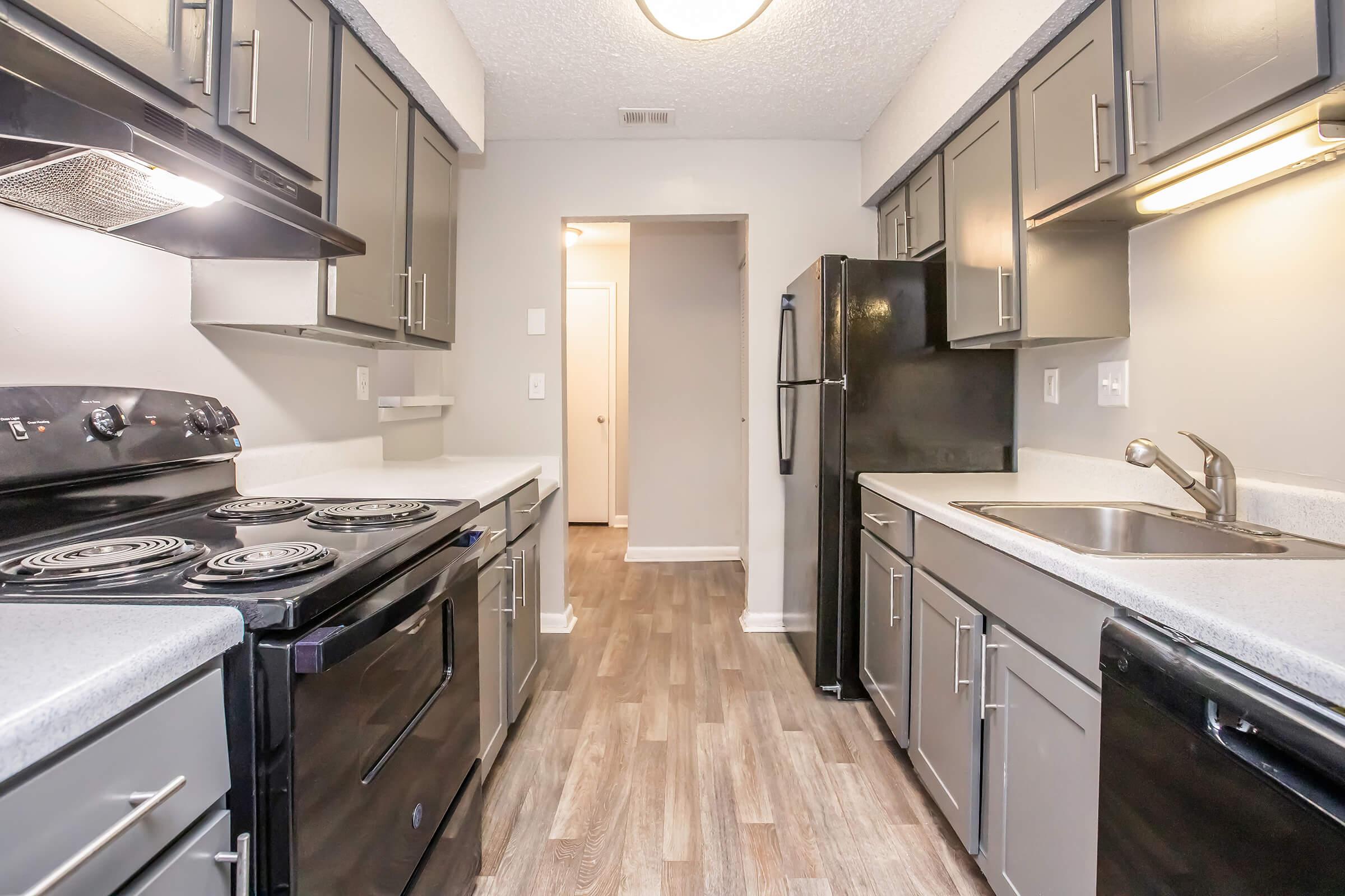 A modern kitchen featuring a gray color scheme with dark cabinets, a black stove, and refrigerator. The countertops are light gray, and the flooring is wood-look laminate. The kitchen has overhead lighting, with an open entryway leading into another room.