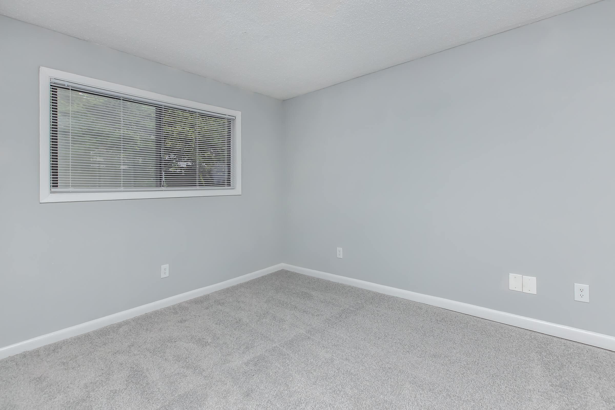A vacant room with light gray walls and carpeted flooring. There is a window with white blinds on the left side, allowing natural light to enter. The room appears bright and spacious, with no furniture or decorations present.