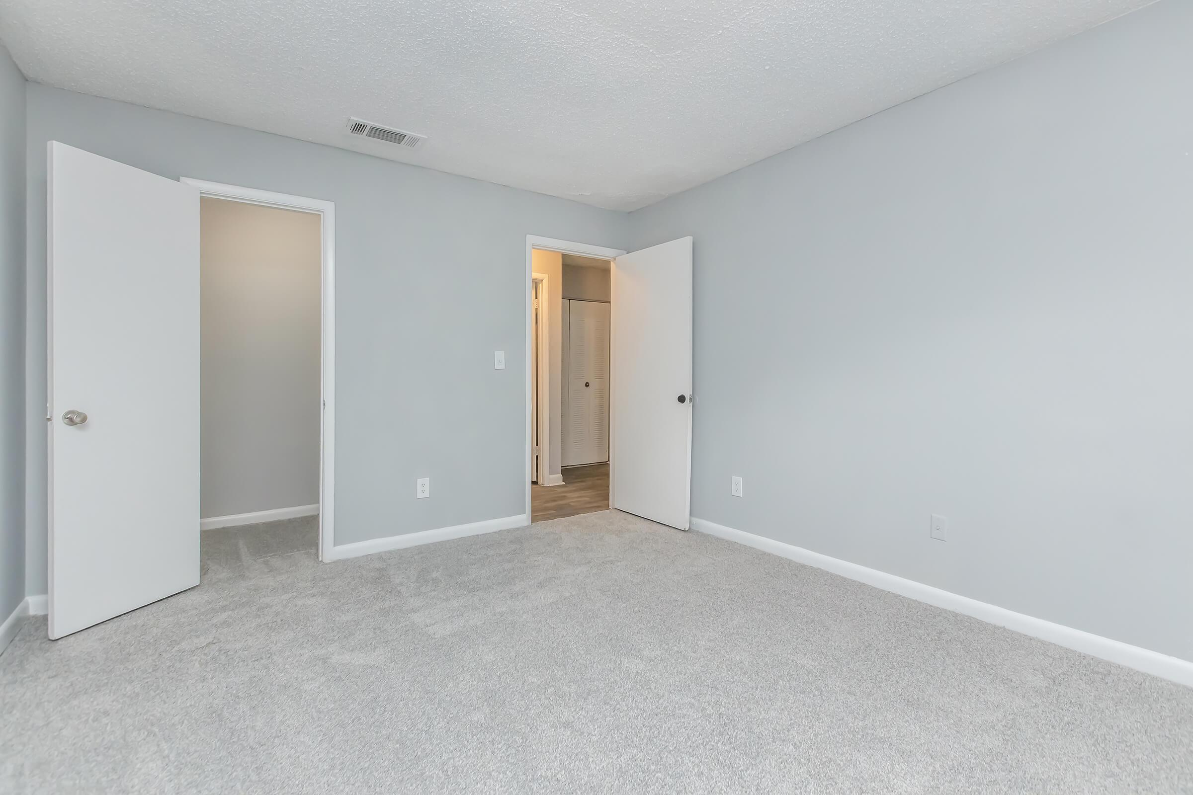 A light gray room with freshly painted walls and new carpet. Two doors are visible on the left, one leading to a closet and the other to an exterior hallway. The room appears empty and spacious, with natural light coming from a doorway at the far end.