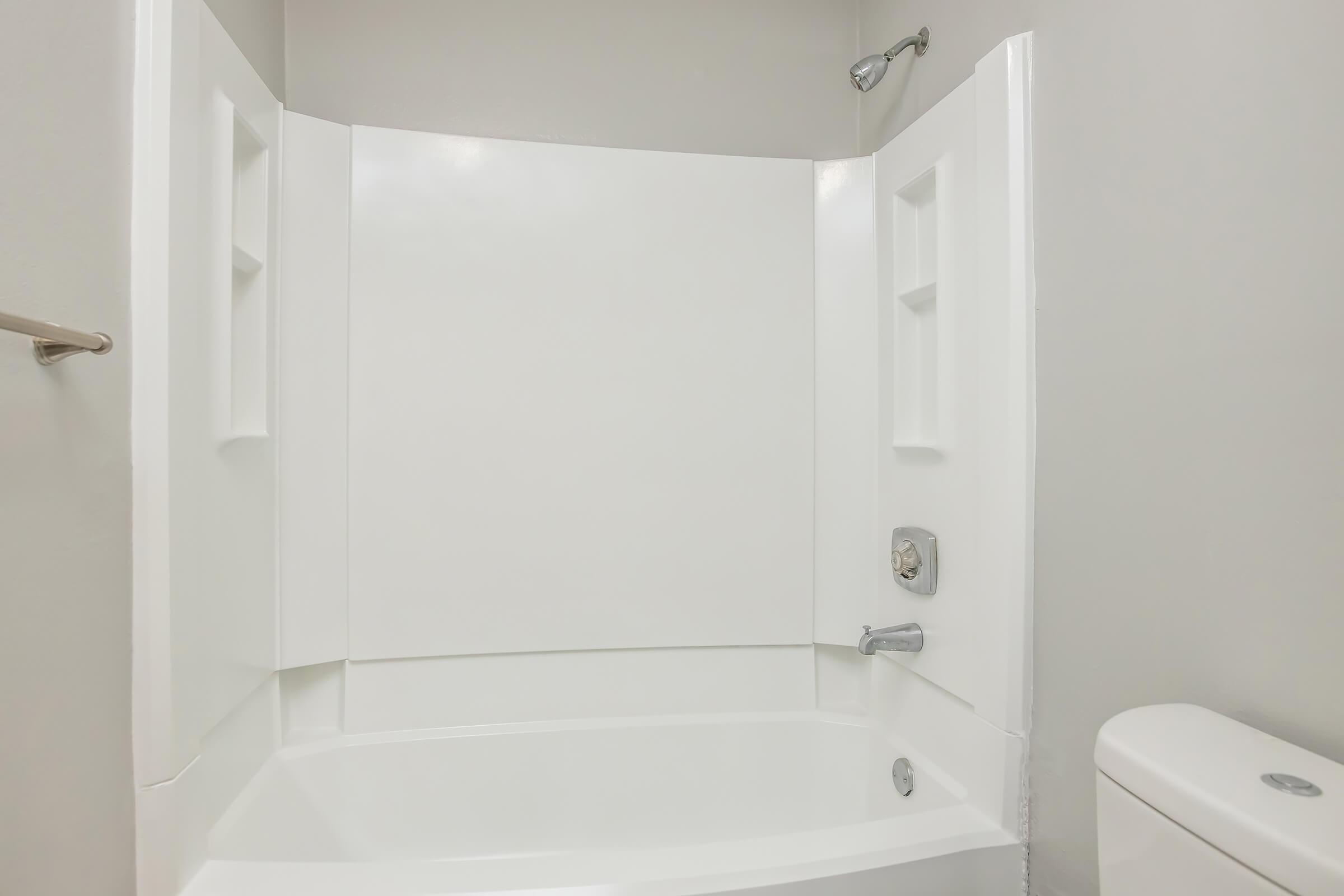 A clean and simple bathroom featuring a white shower/bathtub combination with a wall-mounted showerhead. The background is plain and neutral-colored, emphasizing the minimalist design. A toilet is partially visible on the right side of the image.