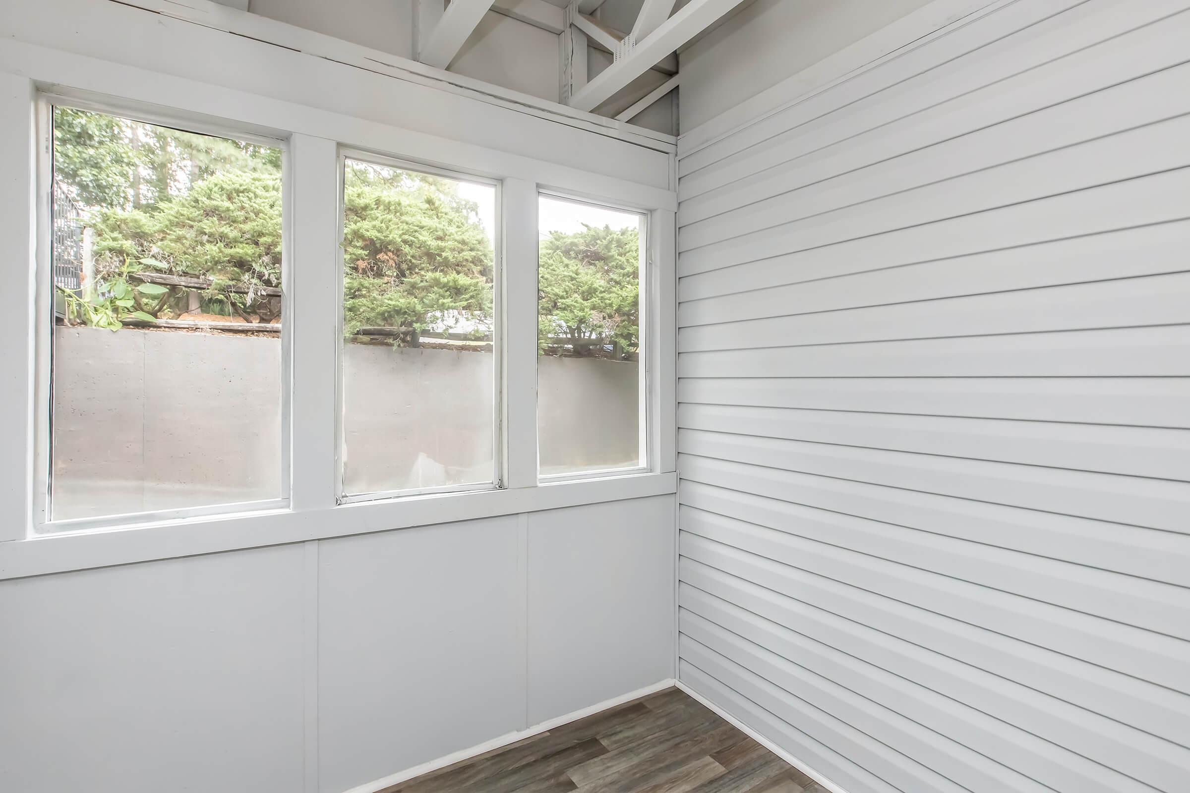 Bright, airy room with white walls and large windows offering a view of greenery outside. The floor features laminate planks, and the ceiling has exposed beams, creating a modern, open feel. The space appears clean and well-maintained, suitable for various uses like a sunroom or leisure area.