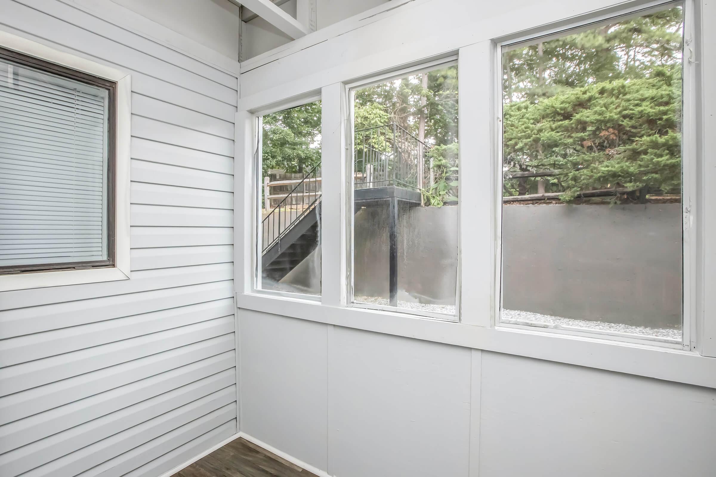A sunroom with large windows overlooking a green outdoor area. The walls are white, and the flooring is wooden. A staircase is visible in the background, leading to an outside space. Natural light fills the room, creating a bright and airy atmosphere.