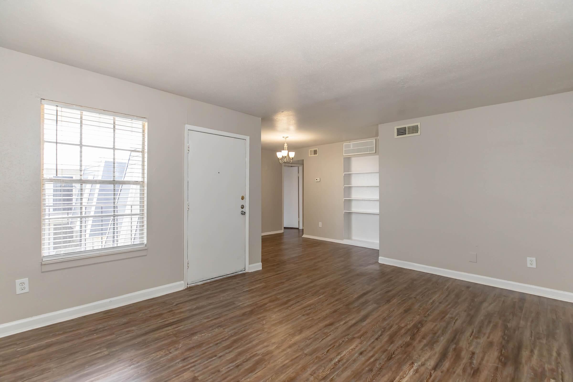 A spacious living area with light-colored walls and wood-style flooring. A window with blinds allows natural light to enter, and an adjacent door leads outside. On the right, there is a built-in shelf, and a doorway in the background leads to another room. The overall ambiance is bright and inviting.