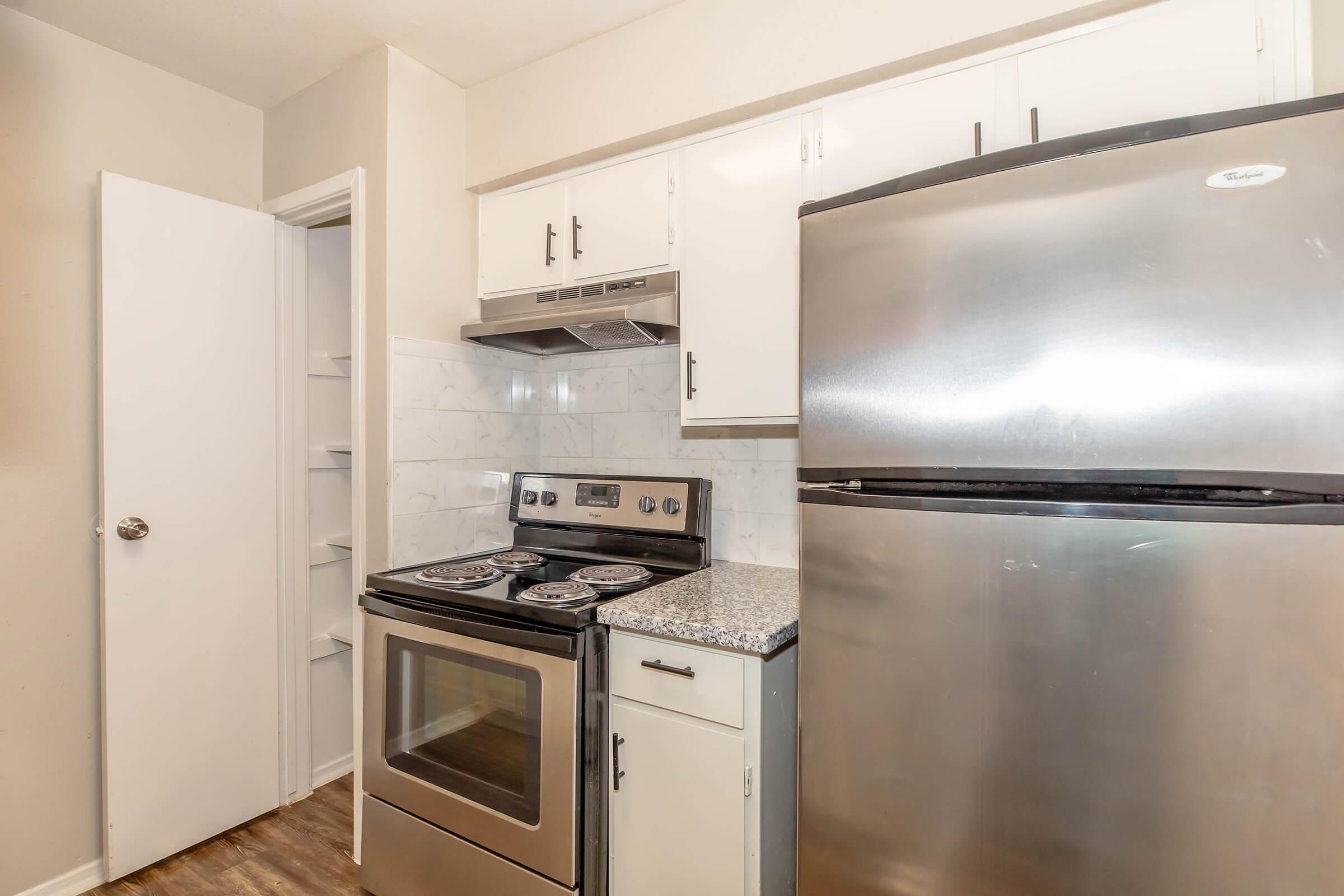 Kitchen featuring a stainless steel refrigerator and stove with a hood, white cabinets, and a granite countertop. The room has light-colored walls and laminate flooring, with a door leading to a small pantry or closet on the left.