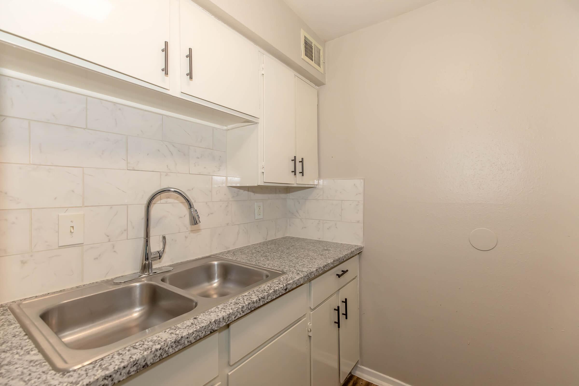 A small kitchen featuring a dual basin stainless steel sink with a modern faucet, white cabinets with black handles, and a light-colored granite countertop. The backsplash is composed of light marble tiles, and the walls are painted a soft neutral color.