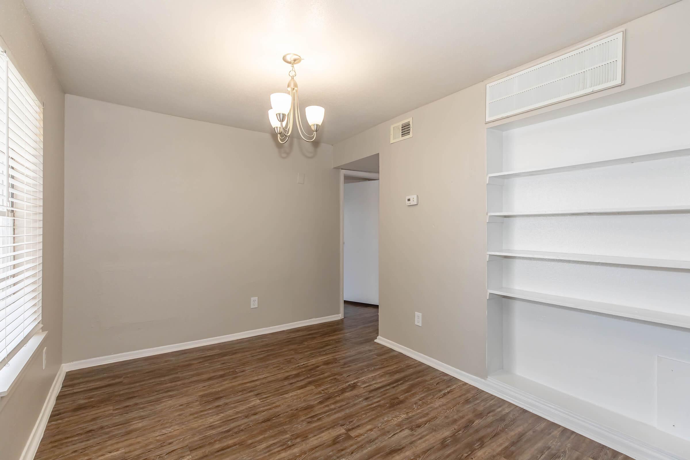 A well-lit, empty room featuring wood-like flooring, light gray walls, and a modern chandelier. There is a set of shelves on the right side and a doorway leading to another room in the back. The window has white blinds, allowing natural light to fill the space.
