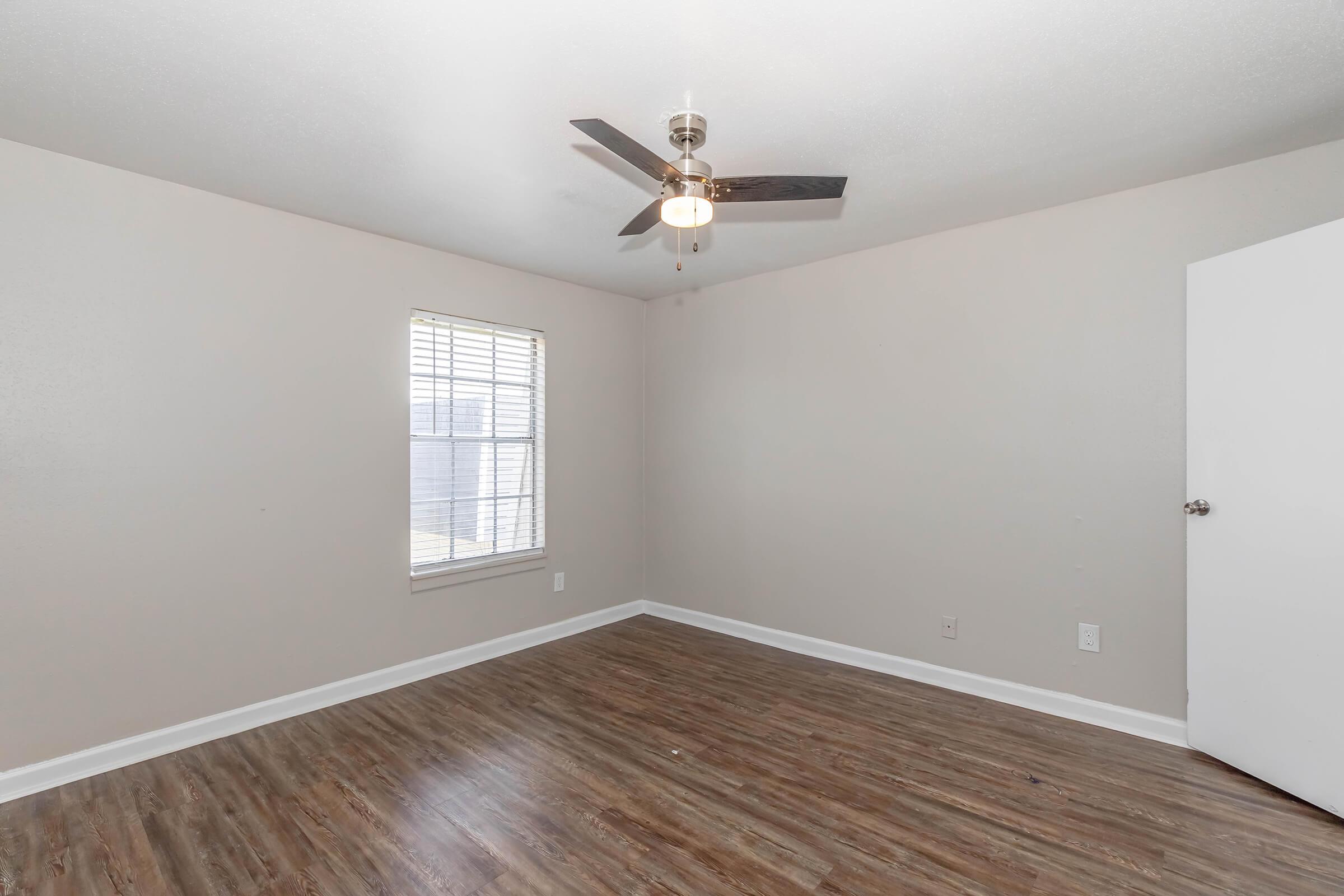 Empty room with light wood laminate flooring, a ceiling fan with lights, and a window on one wall. The walls are painted a soft beige color, and there is a white door on the right side. Natural light is coming in through the window, creating a bright and airy atmosphere.