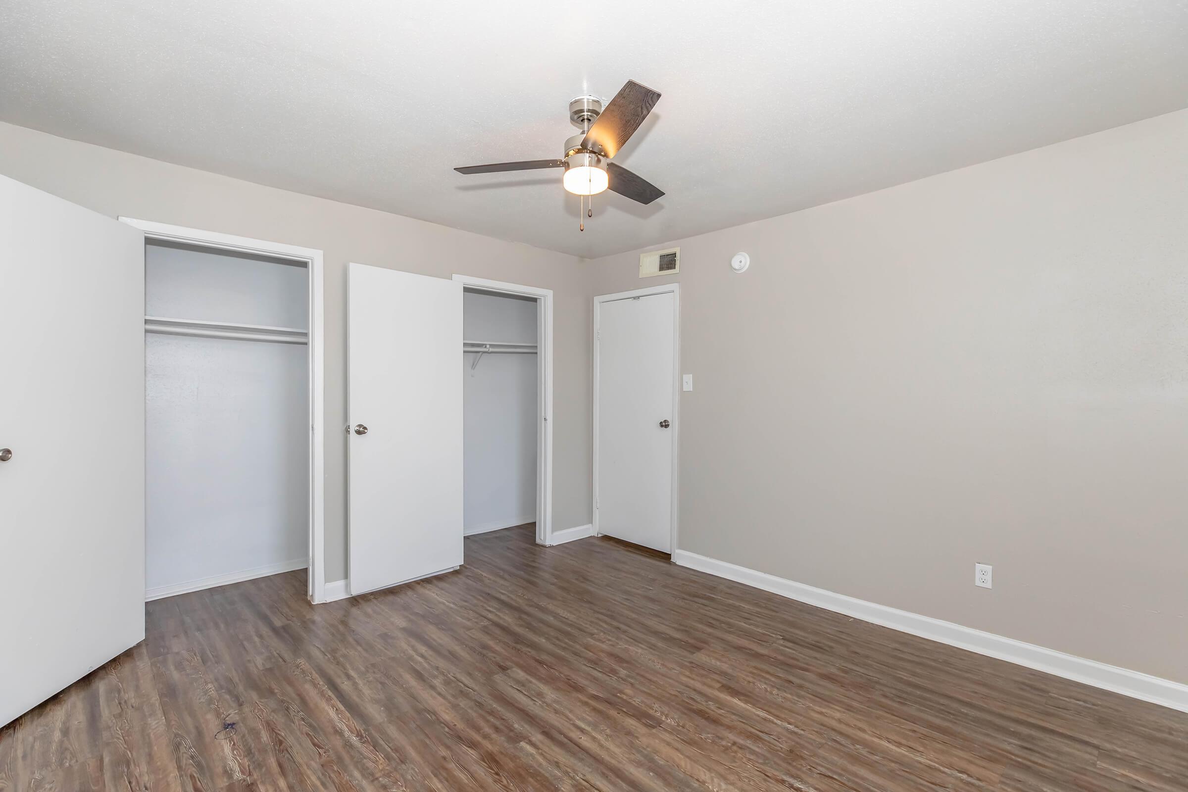 An empty room featuring light-colored walls, a ceiling fan with three lights, and two open closet doors. The floor is a medium brown wood-look laminate. A third door is visible on the far wall, leading to another space. The overall atmosphere is bright and clean.