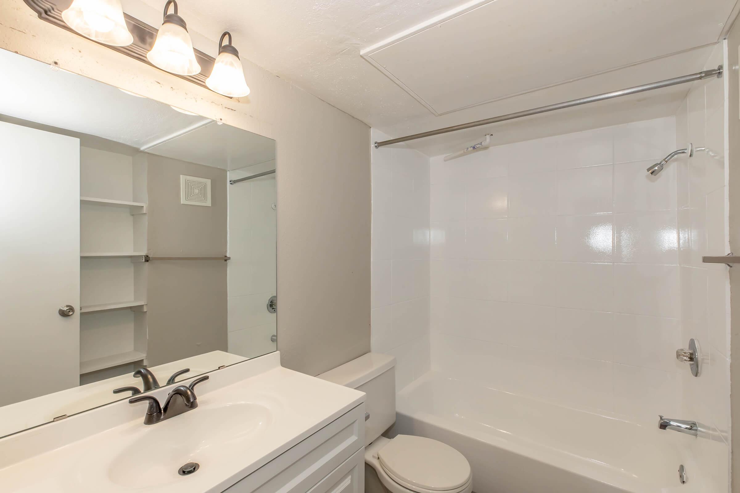 Bright and clean bathroom featuring a white bathtub and shower, a modern sink with a sleek faucet, and a large mirror. The space has light-colored walls, a shelf unit for storage, and stylish lighting above the mirror, creating a fresh and inviting atmosphere.