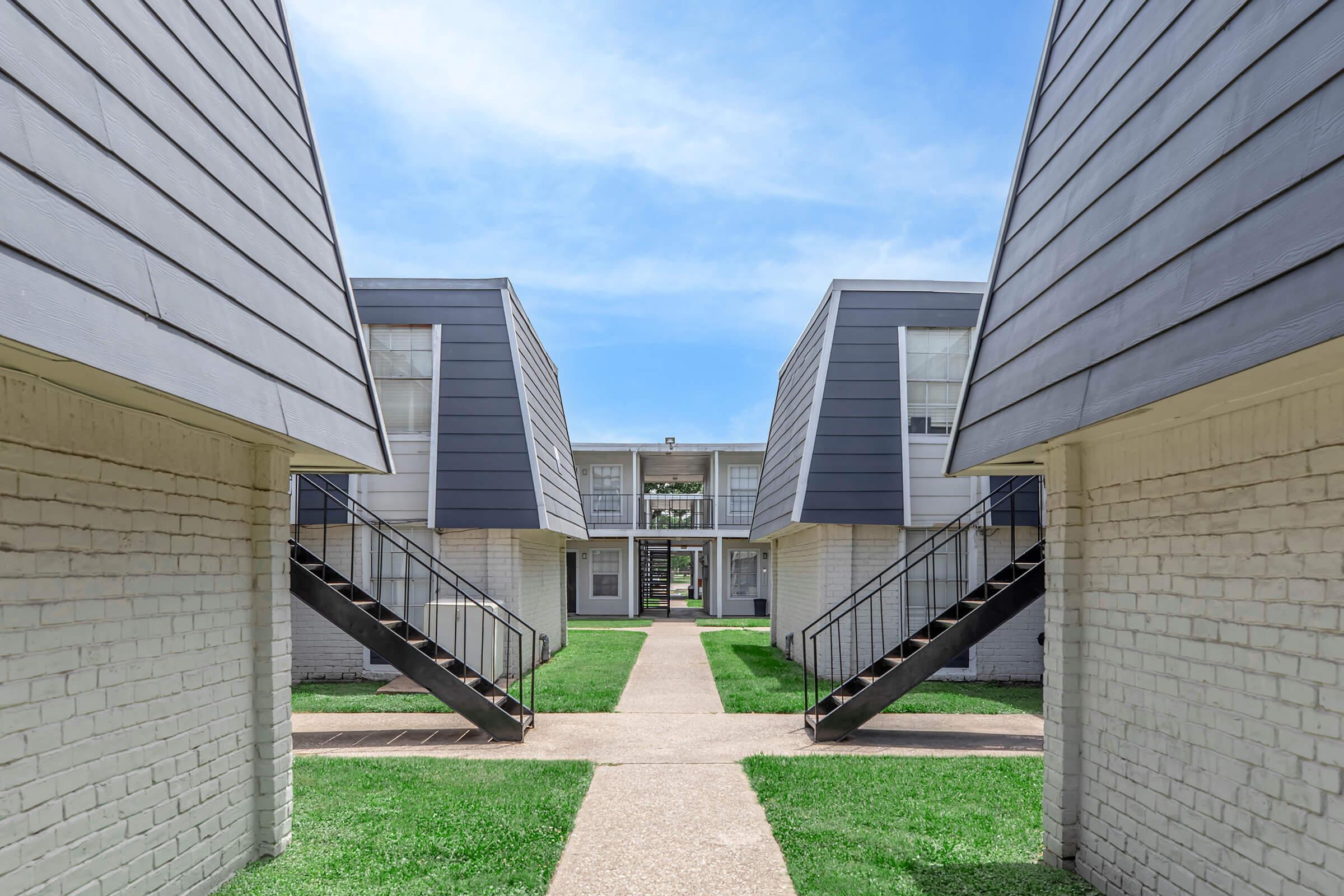 View of a residential complex featuring two buildings with sloped roofs, separated by a grassy pathway. Each building has a set of stairs leading to upper levels, and the scene is illuminated by a clear blue sky, creating a bright and inviting atmosphere.