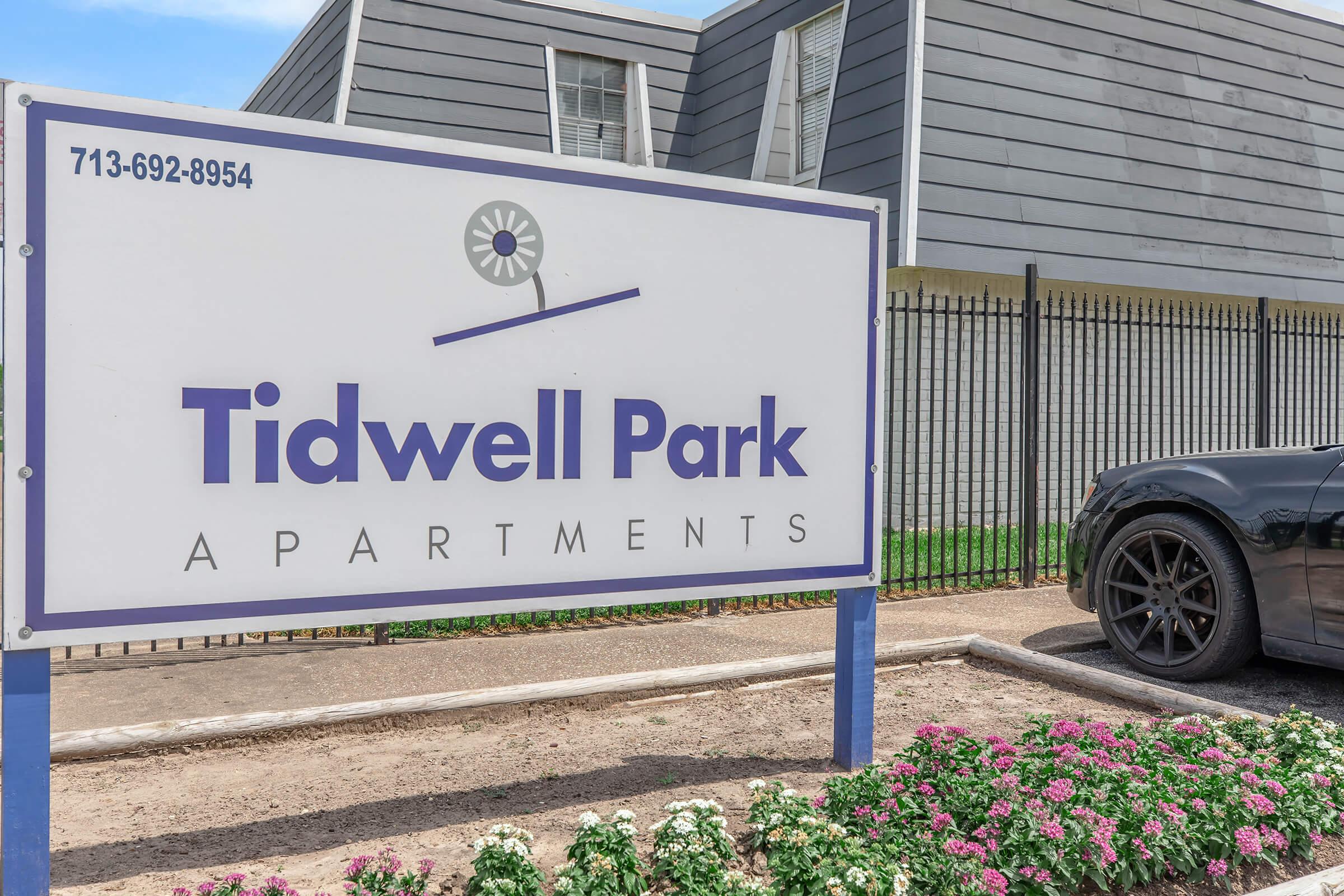 Sign for Tidwell Park Apartments, featuring the name prominently displayed in purple text against a white background. A patterned border surrounds the sign, with a nearby black car parked beside a flower bed of colorful flowers. In the background, a modern gray building can be seen.