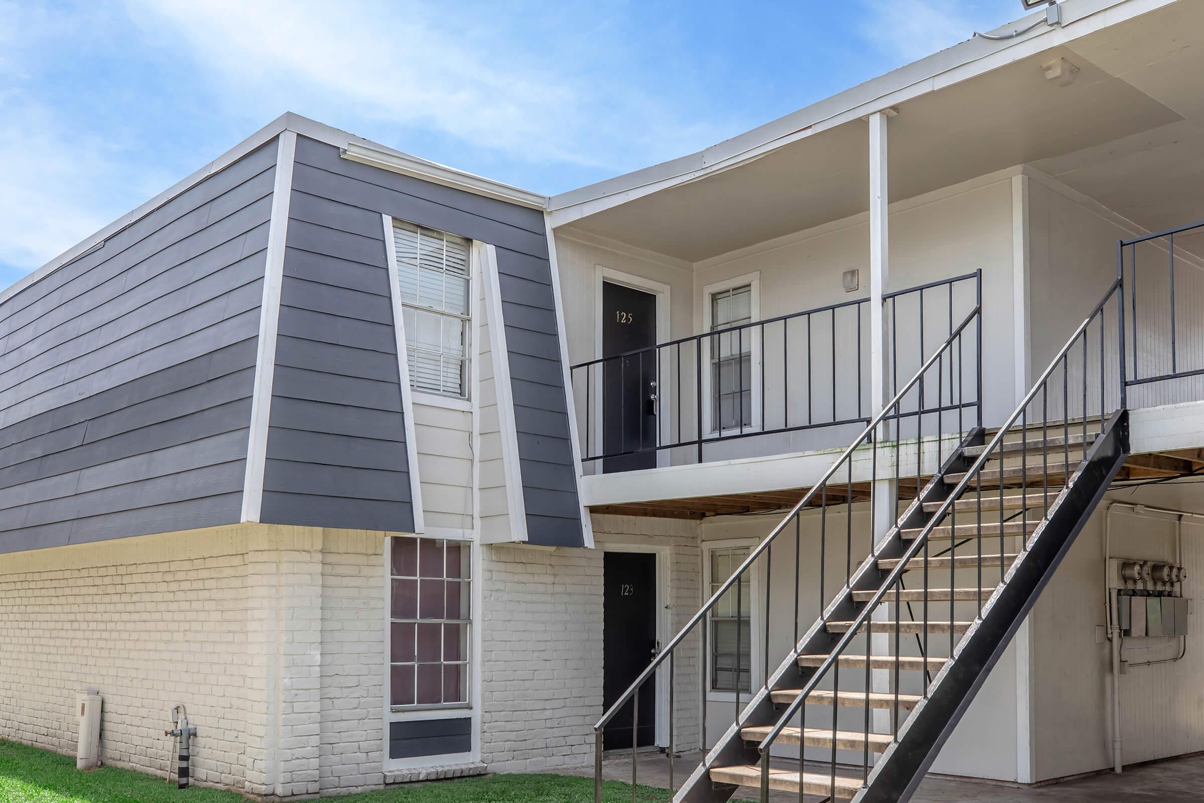 A modern apartment building with a unique, angled exterior design. The structure features a combination of gray siding and white brick, with a staircase leading to the upper level. The entrance doors are numbered, and the grassy area in front complements the architectural style.