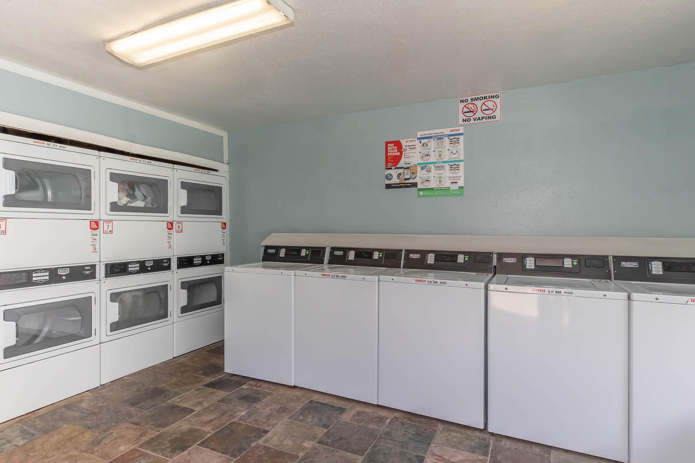 A clean laundry room featuring several stacked dryers on the left and row of washing machines on the right. The walls are painted light blue and there's a tiled floor. A sign with laundry rules is posted on the wall, and overhead lighting provides bright illumination.
