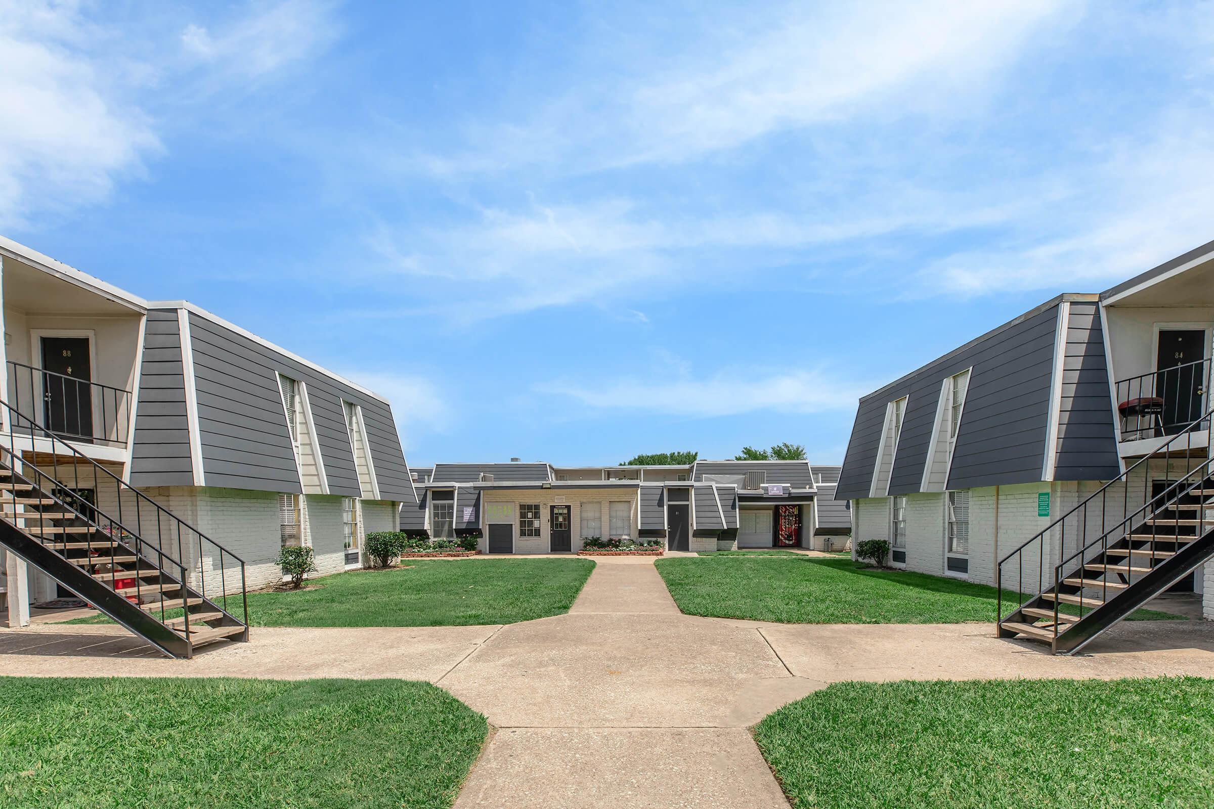 A view of a residential apartment complex featuring two-story buildings with gray sloped roofs, surrounded by green grass. A central pathway leads through the courtyard, connecting to various entrances, with stairs on either side. The sky is partly cloudy, indicating a bright day.
