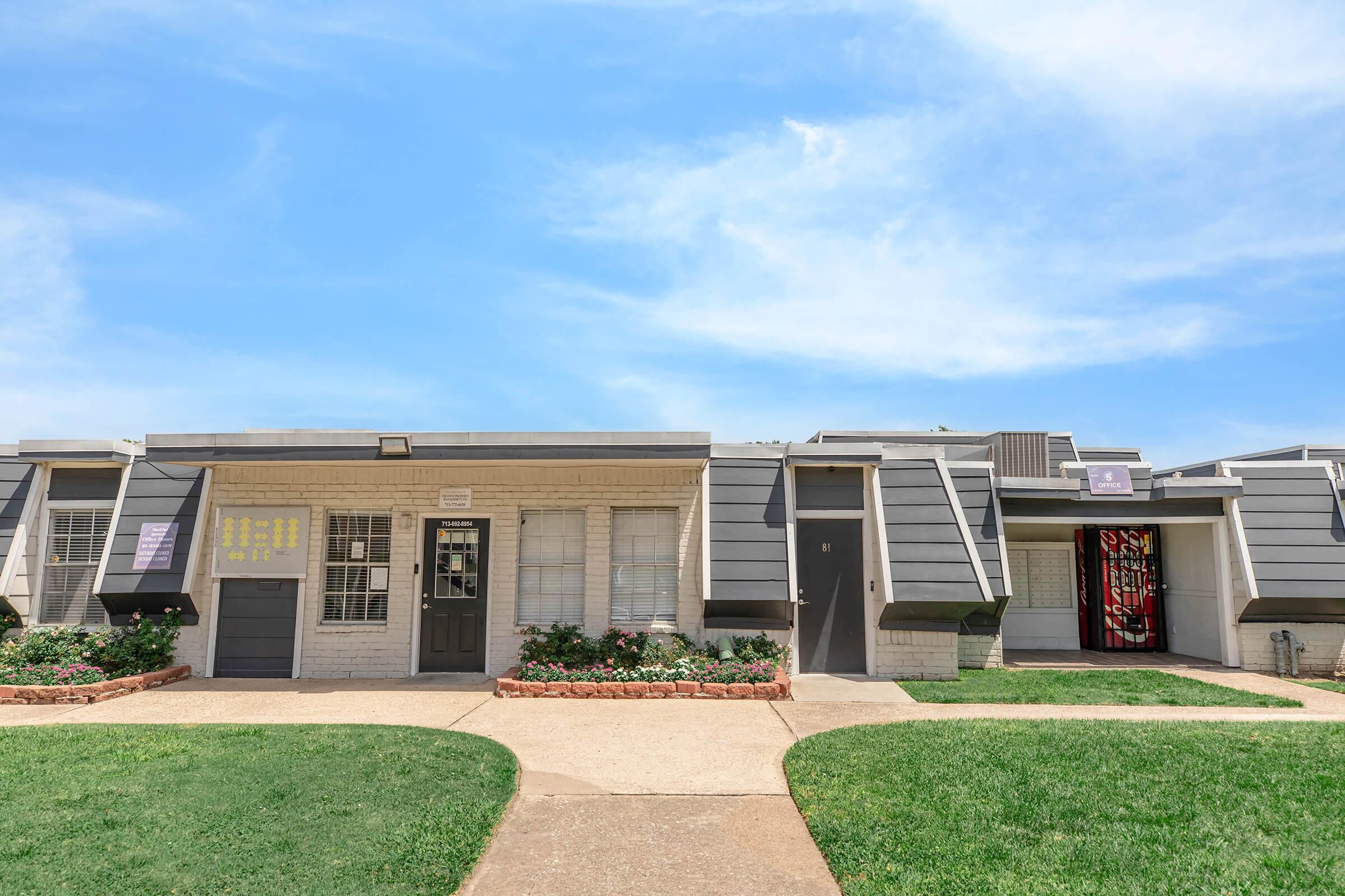 Two-story apartment buildings with gray exteriors and dark doors, surrounded by green grass and flower beds. The façade features a mix of windows and entrance ways, with some units displaying signs. A clear blue sky provides a vibrant backdrop to the scene.