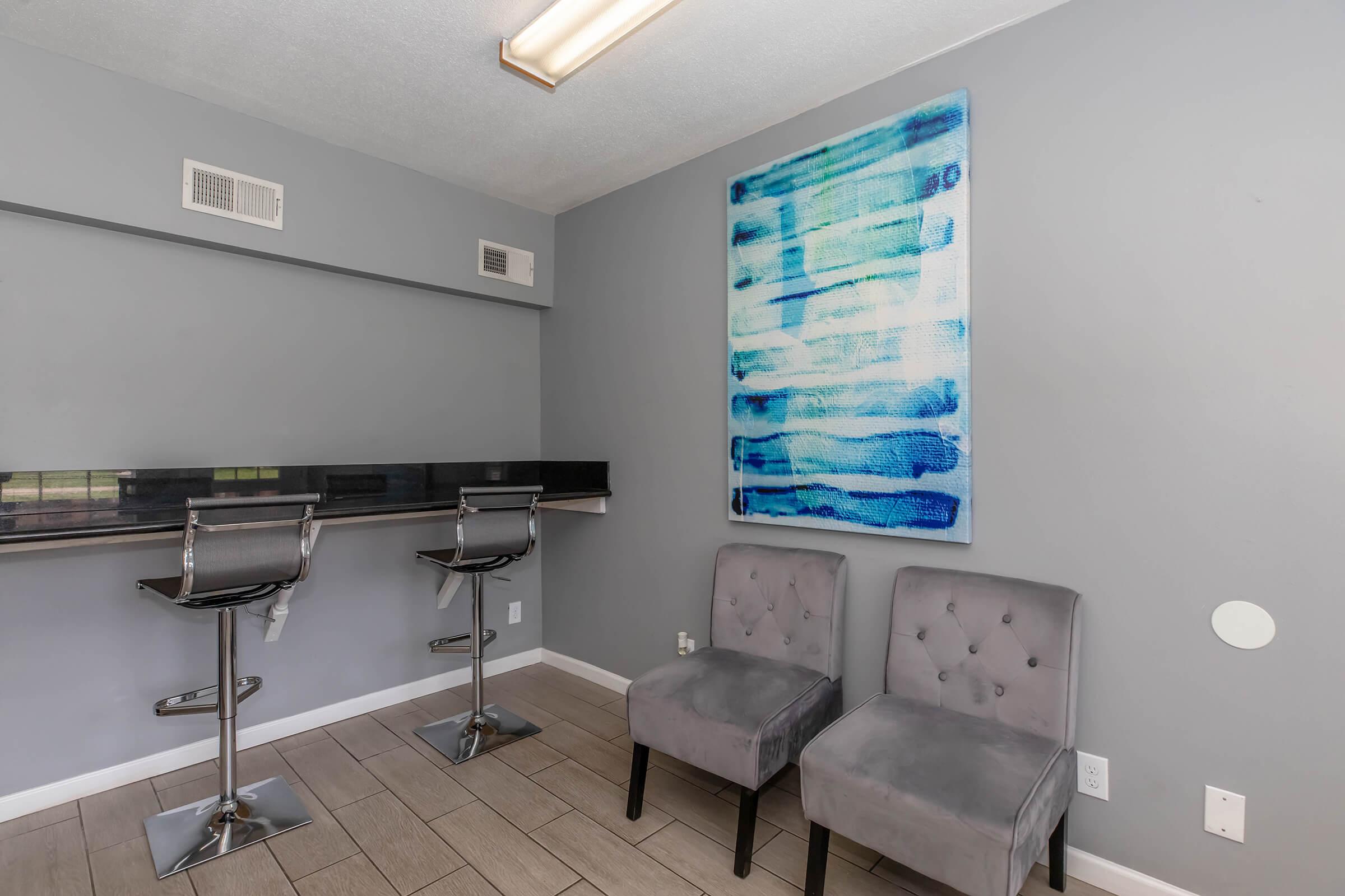 A modern interior space featuring a gray wall, a colorful abstract painting, two stylish gray chairs, and a bar area with two black barstools. The flooring is light-colored tile, and the overall decor has a contemporary aesthetic.