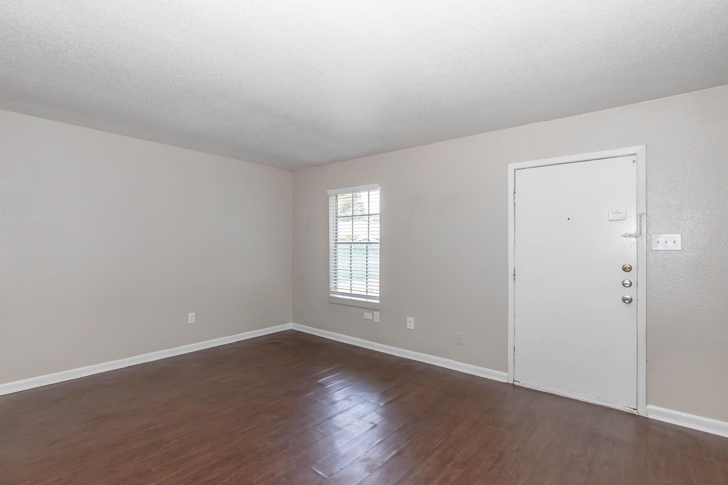 Empty room with light-colored walls and a dark wood floor. A window with white blinds allows natural light, and a plain door is visible on the right. The space is unadorned and features a simple design.