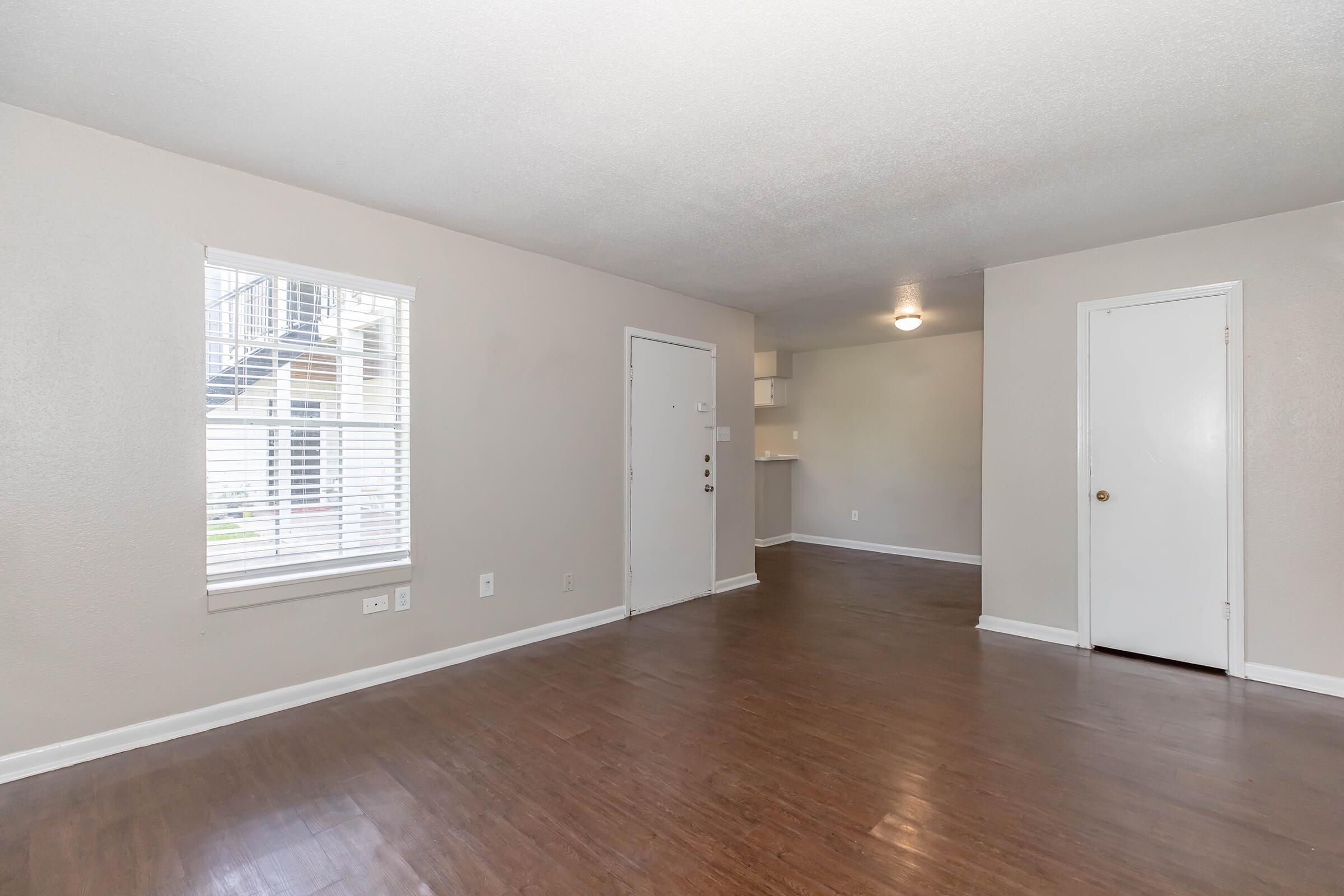 A spacious interior with light-colored walls and a wooden floor. There is a window with white blinds, a front door, and an adjacent room with a small kitchen area visible in the background. The overall space is well-lit and uncluttered, creating a welcoming atmosphere.