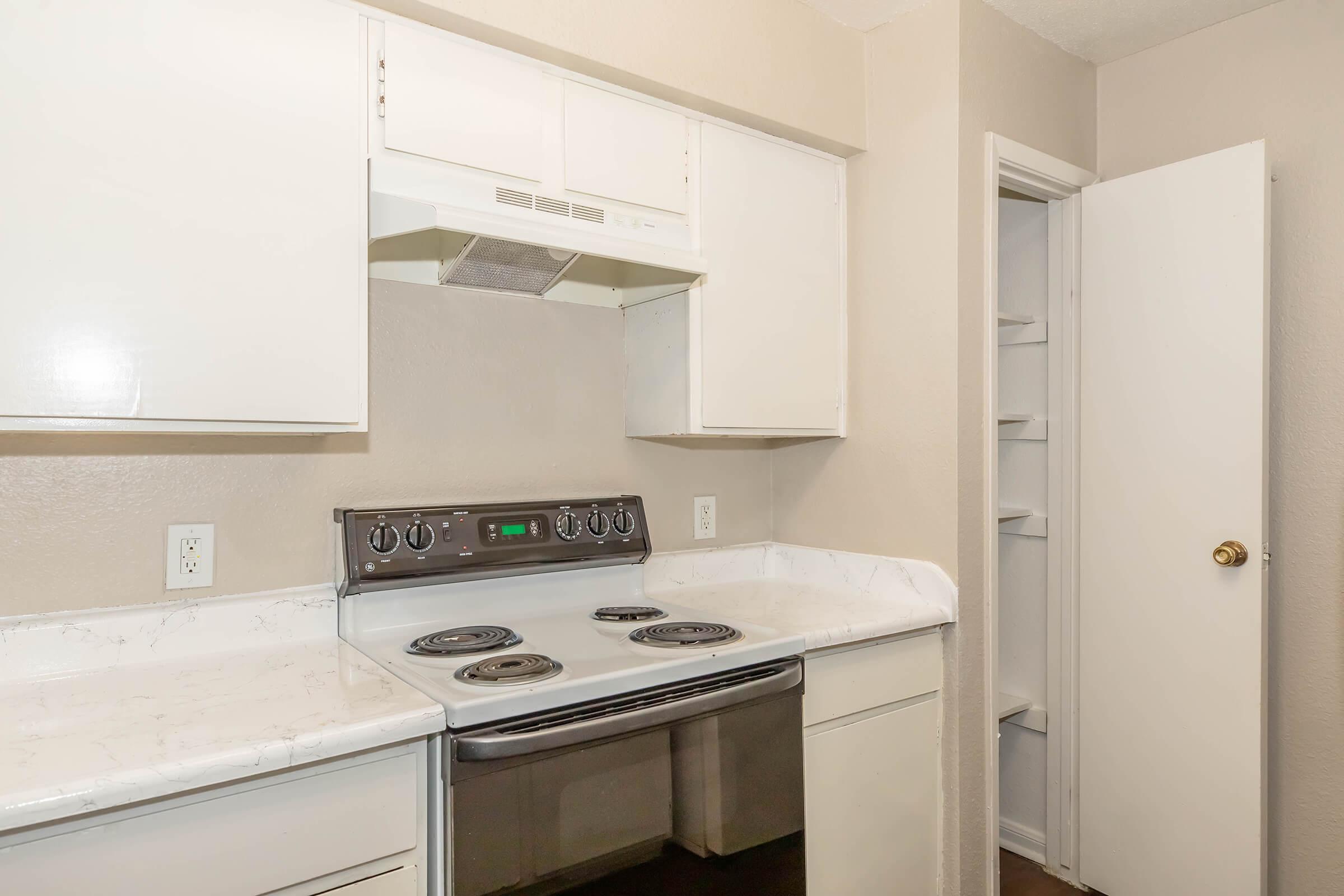 Interior view of a kitchen featuring a white countertop, a stainless steel oven with four burners, an overhead range hood, and white cabinets. A door to a small pantry or storage area is visible on the right, with light beige wall color surrounding the space.