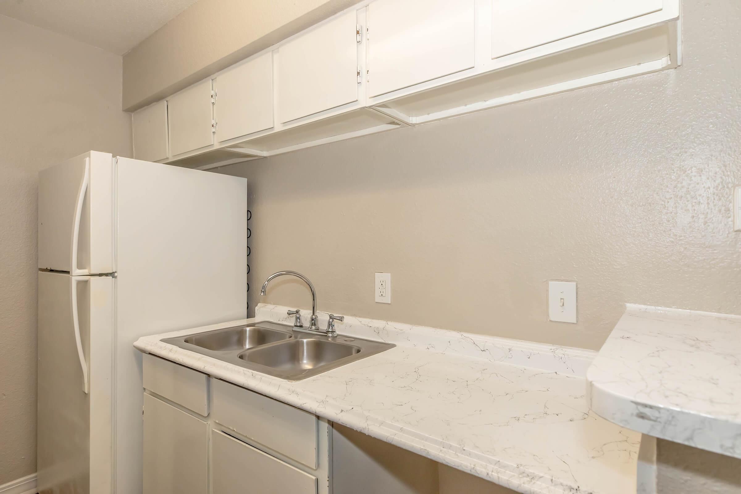 A clean, modern kitchen featuring white cabinetry and a white refrigerator. The countertop is made of light-colored marble, with a double sink visible. The walls are painted in a neutral shade, creating a bright and open atmosphere.