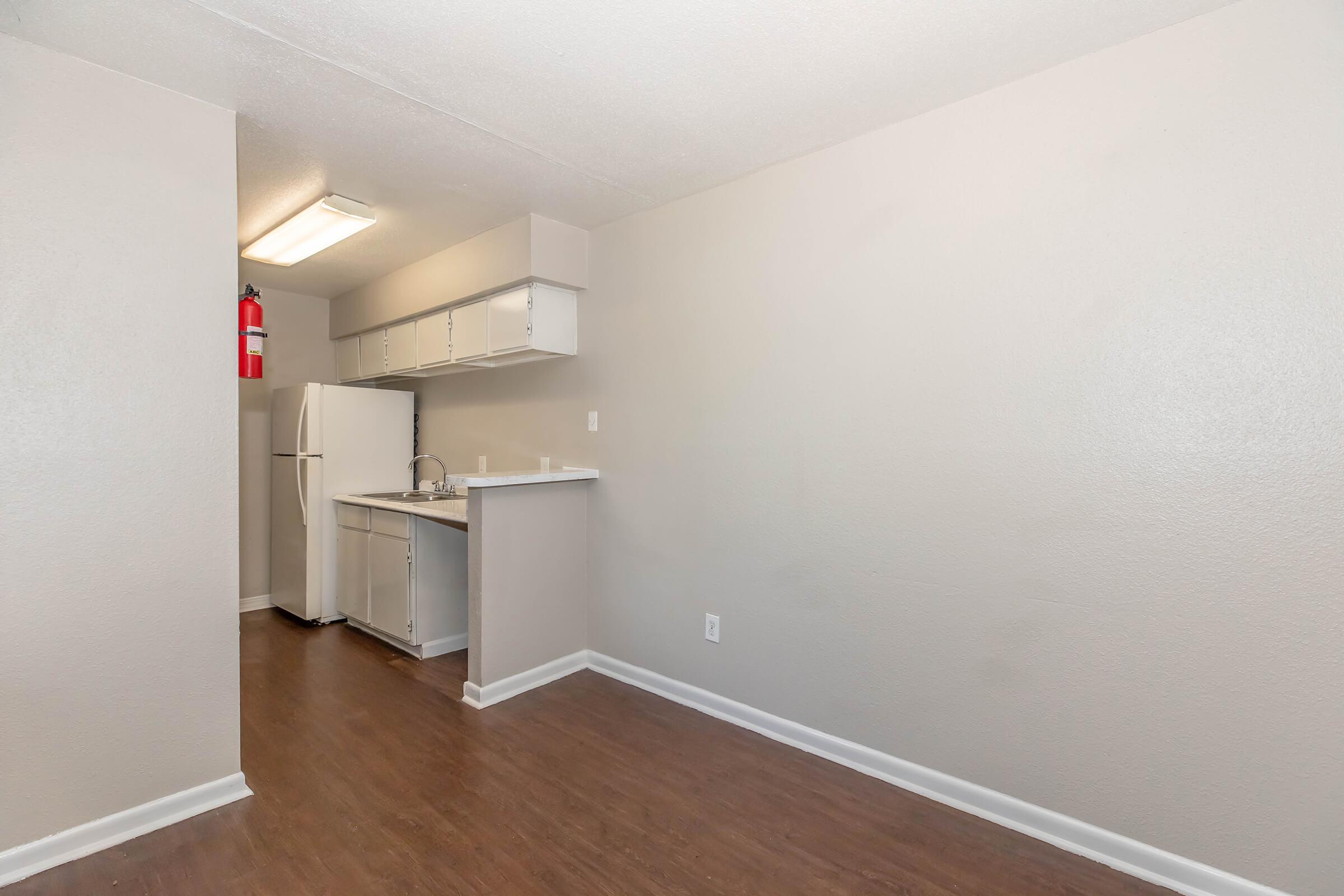 A small, modern kitchen area in an apartment, featuring light beige walls, a white refrigerator, and a countertop with a sink. The flooring is dark wood laminate. Basic cabinetry is present above the counter, and a fire extinguisher is mounted on the wall. Natural light is minimal.