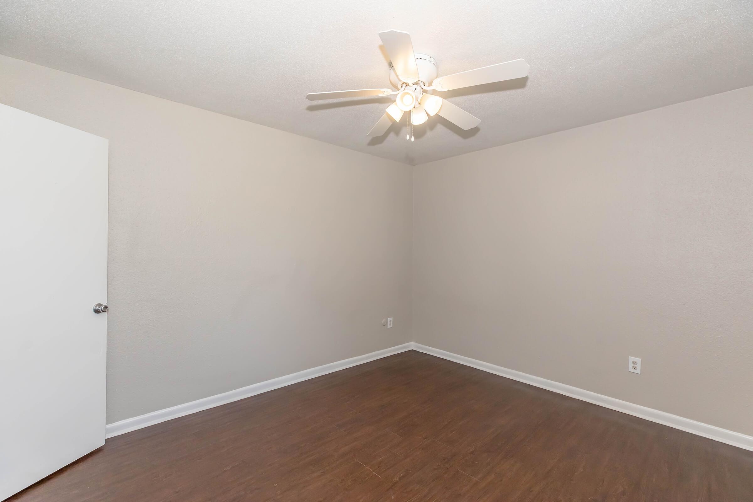 A spacious, empty room with light gray walls and a dark wood floor. There is a white ceiling fan with five blades illuminated by a central light fixture. A closed white door is visible on the left side of the image. The room is well-lit and ready for furnishing.