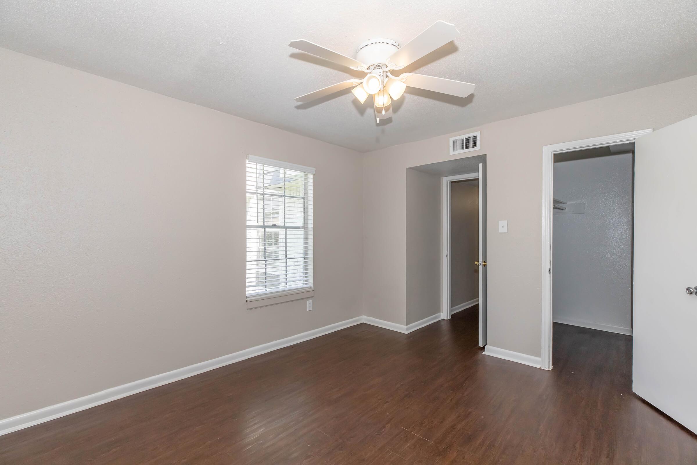 A well-lit room with a ceiling fan, neutral-colored walls, and dark wood flooring. A window with blinds allows natural light, and there's a door leading to another room. The overall space appears clean and inviting, suitable for various furnishings.