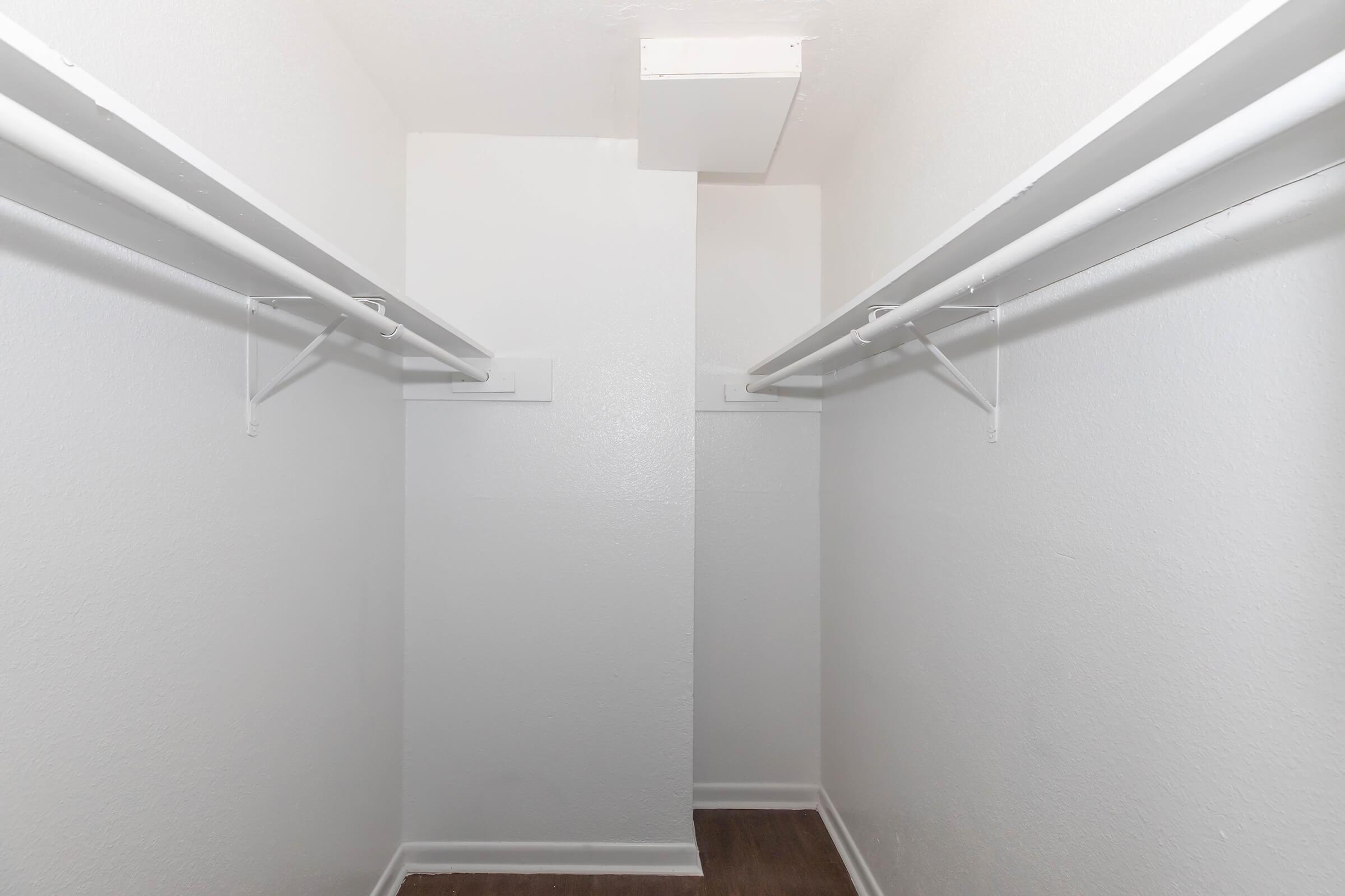 A spacious, empty closet with white walls and two parallel clothing rods. The floor has a light wood finish, and there is an overhead vent visible at the top. The overall look is clean and minimal, providing a blank canvas for storage organization.