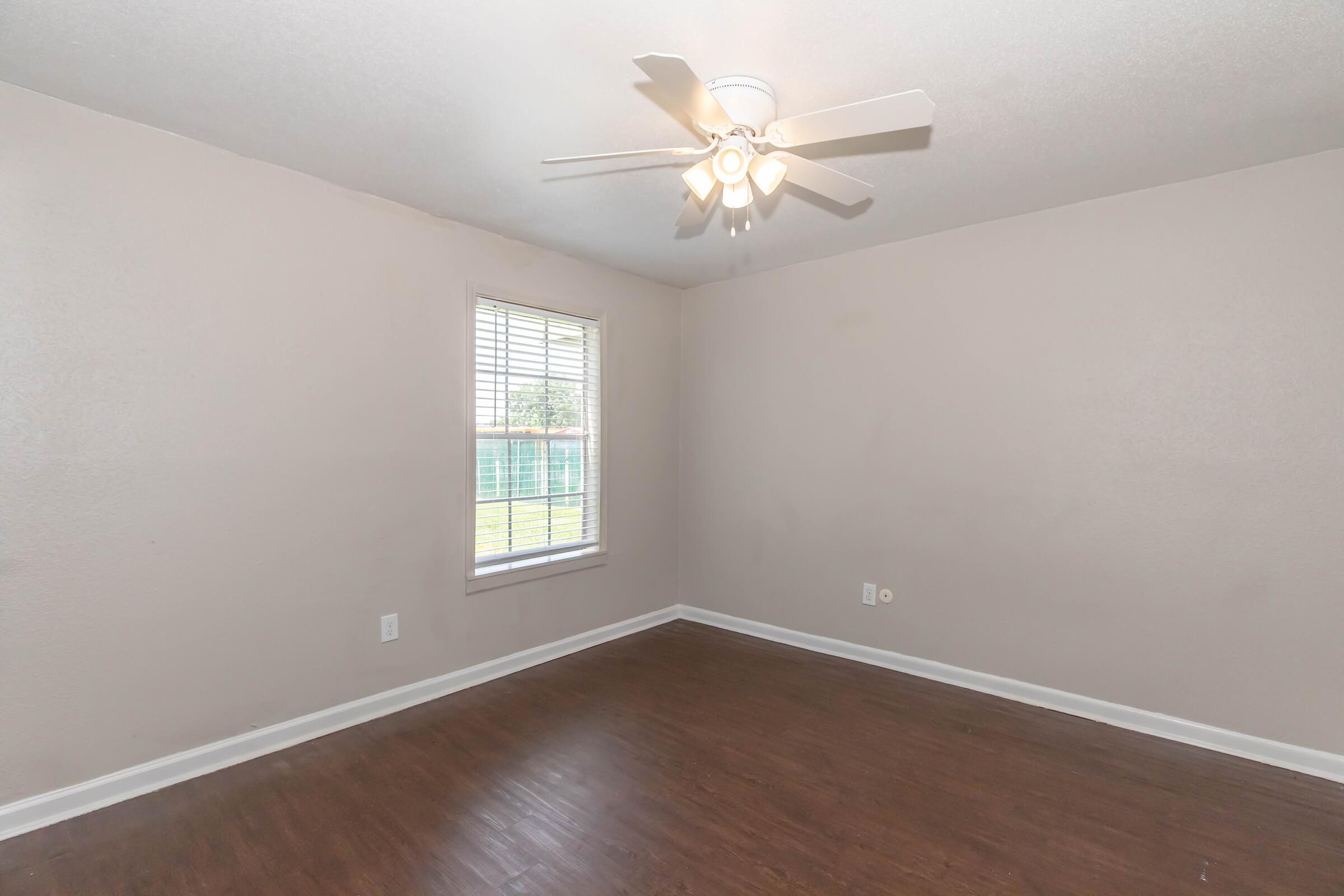 A spacious room with light brown walls and a ceiling fan. The floor is dark wood, and there is a window with white blinds letting in natural light. The space is empty, providing a blank slate for decoration.