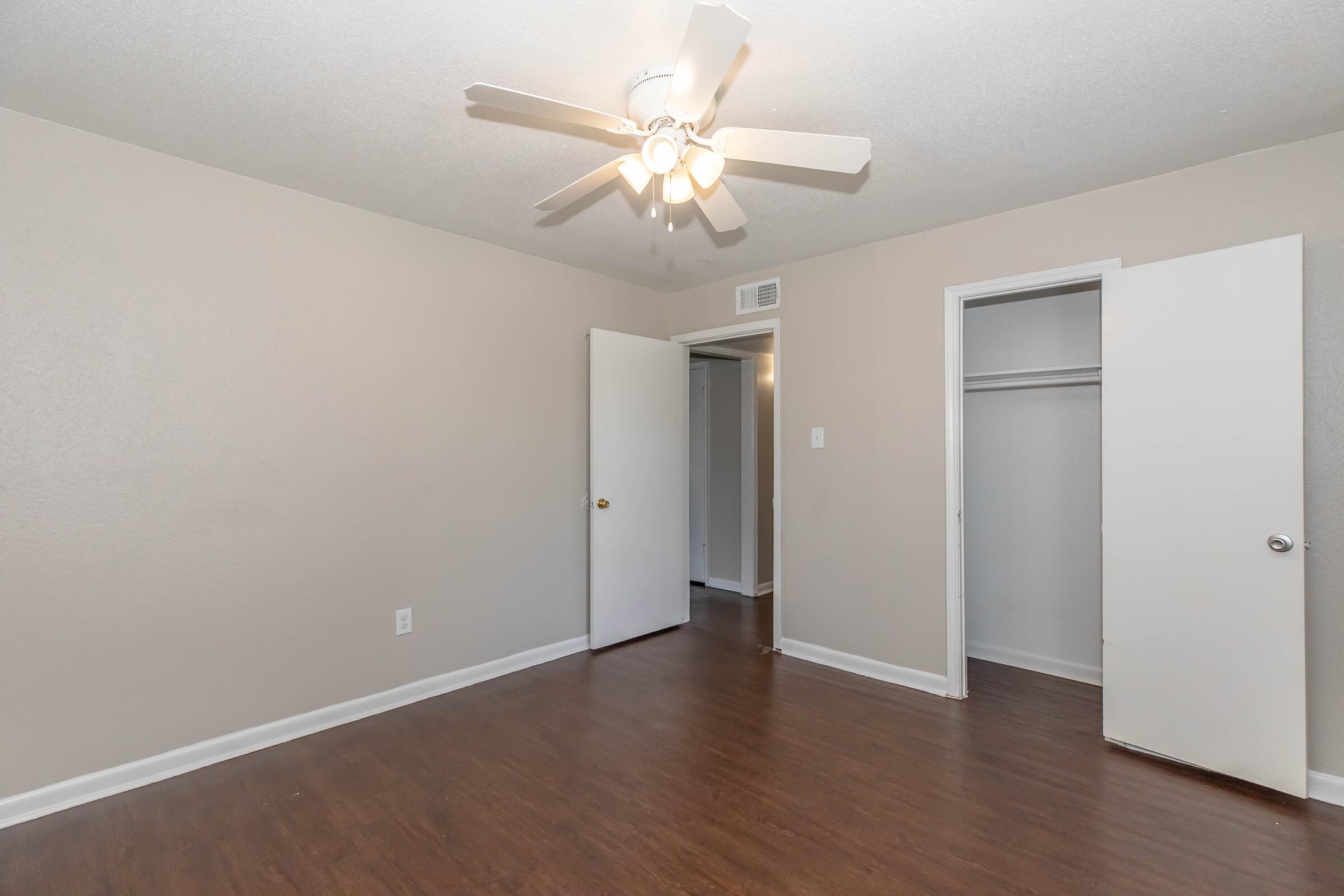 A sparsely furnished bedroom featuring light brown walls, a ceiling fan with six blades, and wood laminate flooring. Two white doors lead to closets, and there is a doorway to another room in the background. The space is well-lit and appears inviting.