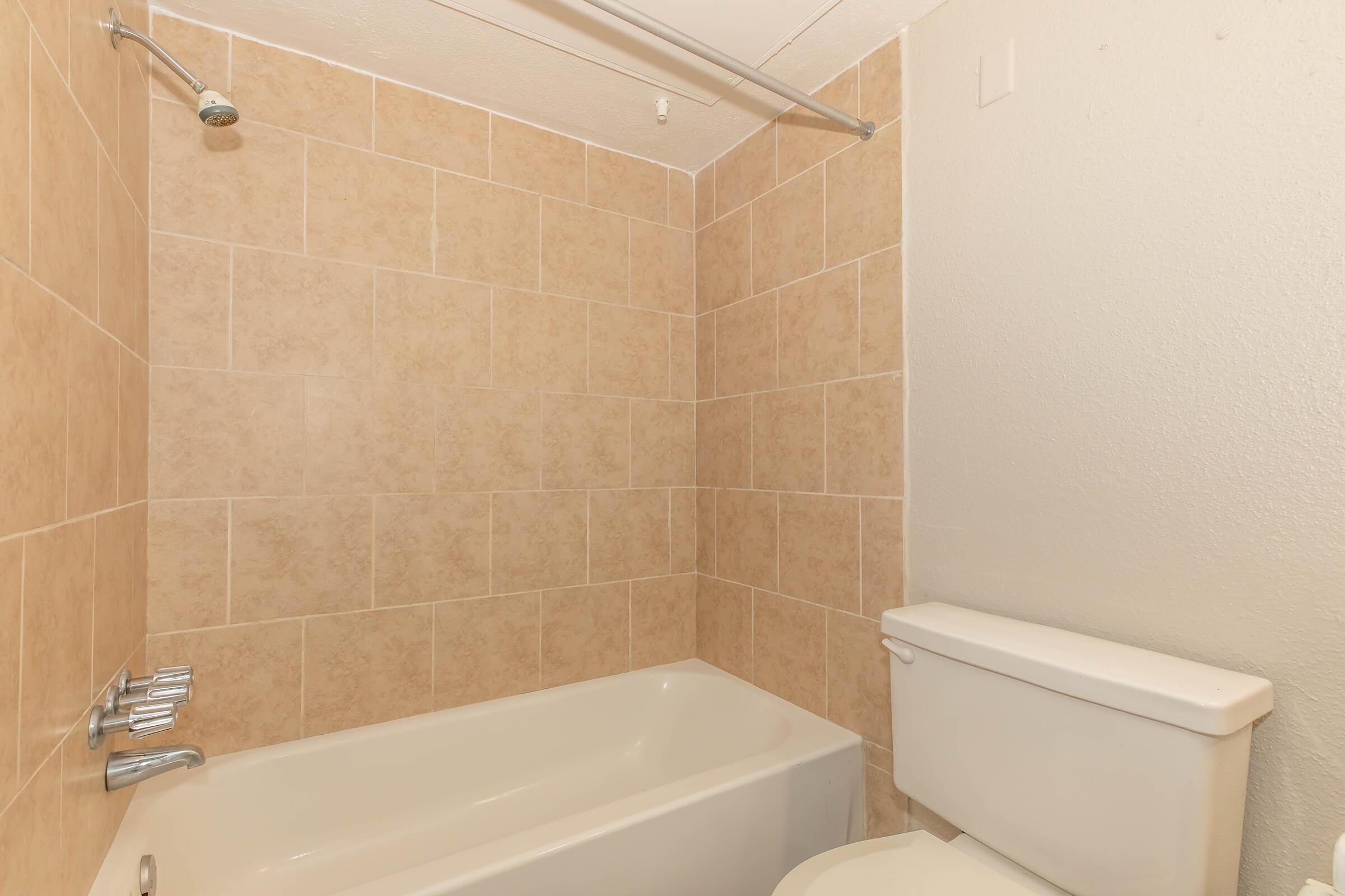 A bathroom featuring light-colored tiled walls, a bathtub with a shower and a silver faucet, and a toilet adjacent to the bathtub. The bathroom has neutral-toned tiles and a simple, clean design.