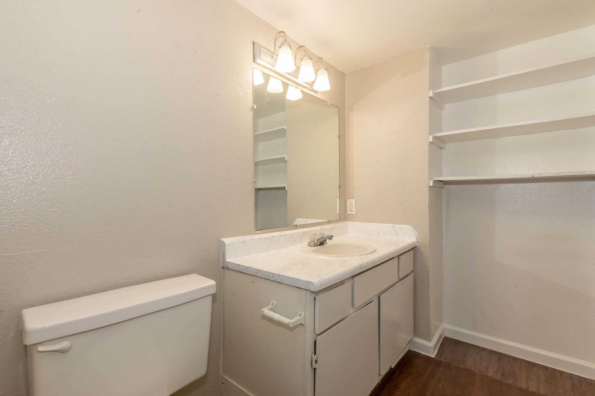 A small, minimalist bathroom featuring a white sink with a countertop, a wall-mounted mirror, and three light fixtures above. There is a white toilet, and to the right, built-in shelves provide storage space. The walls are painted a light color, and the flooring appears to be a dark wood laminate.