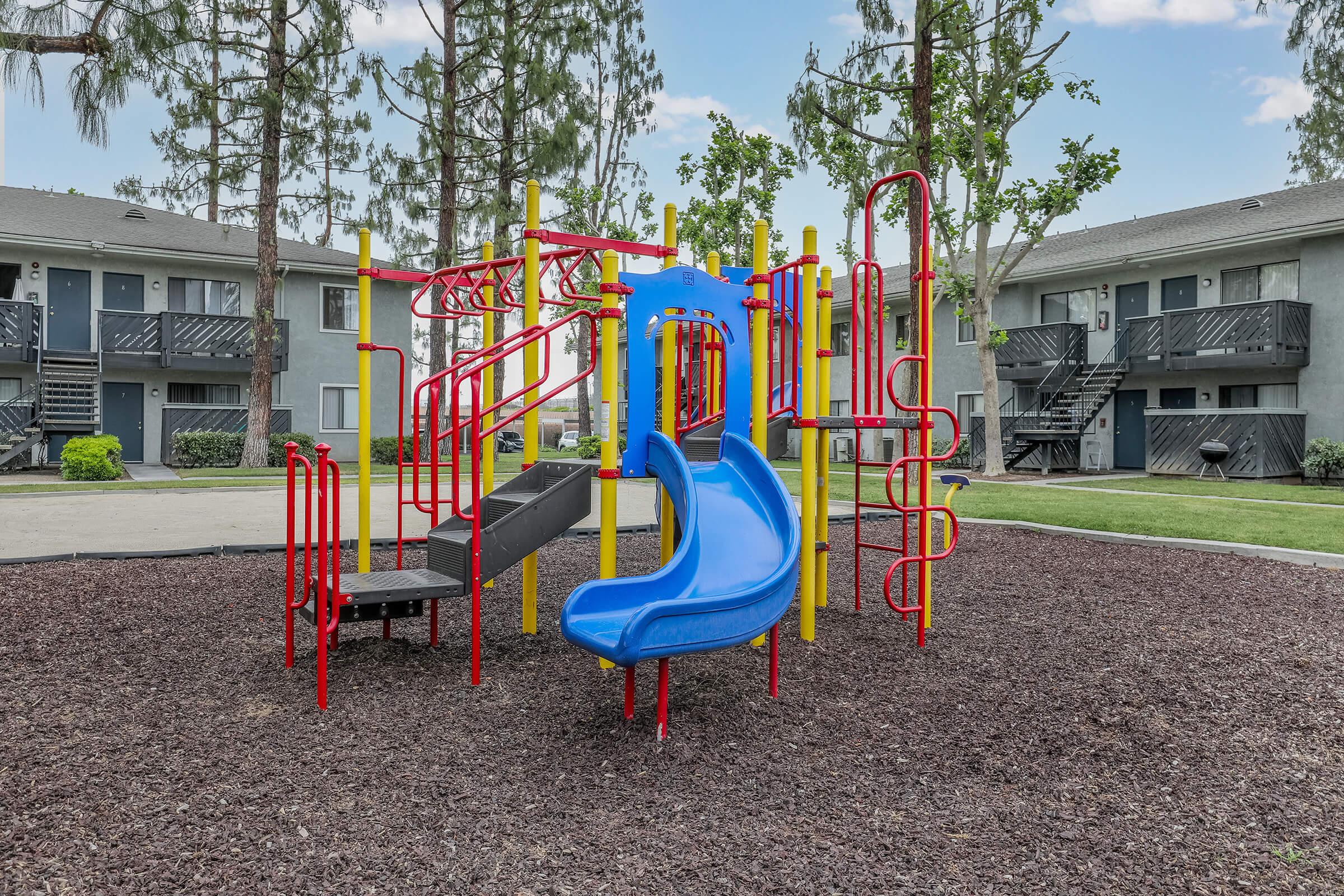 San Bernardino CA Apartments for Rent - Creekside Village - Colorful Playground surrounded by Mulch, landscaped Grounds, and Trees with Apartment Buildings in the Background.