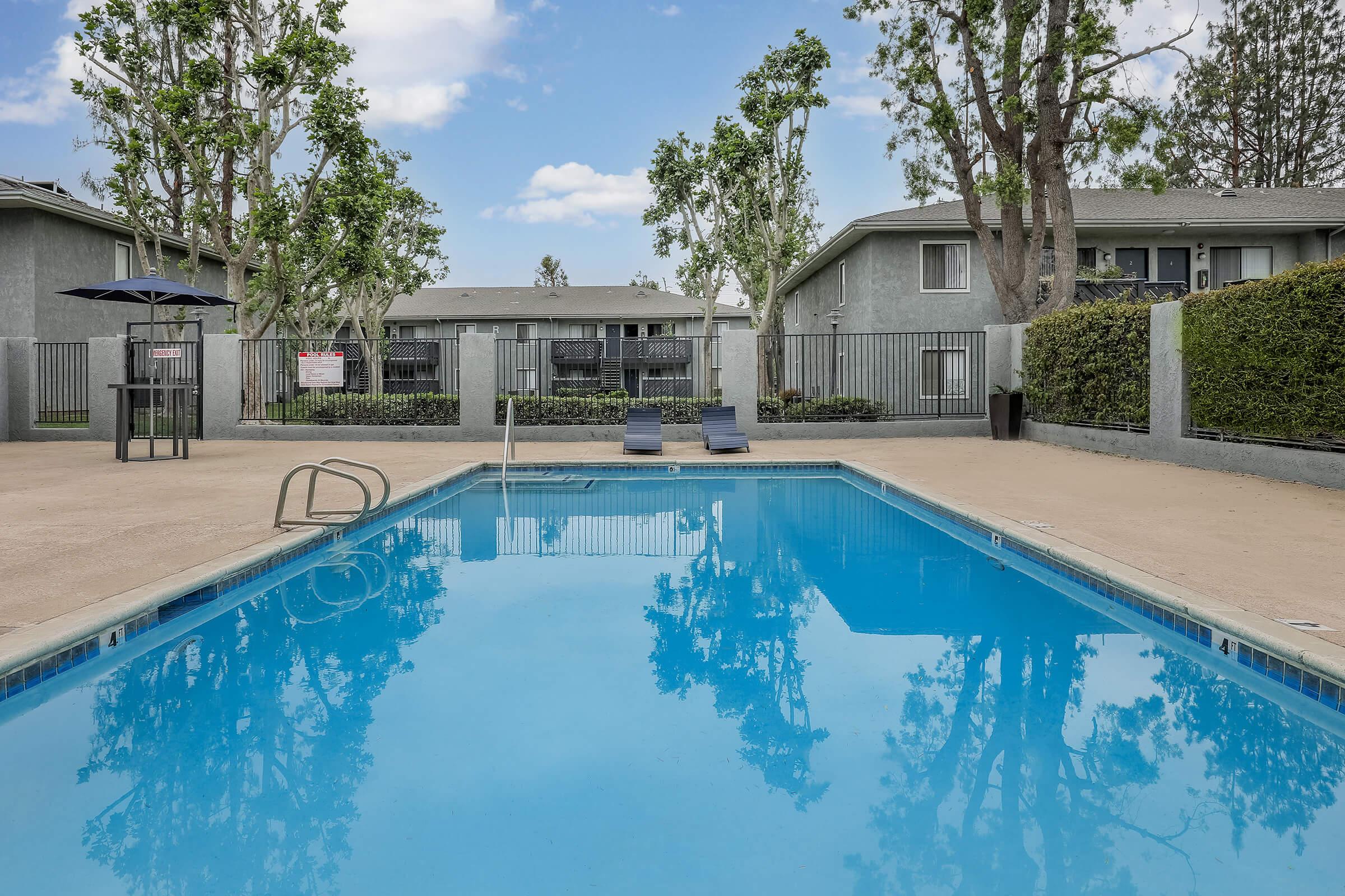 One Bedroom apartments in San Bernardino CA - Creekside Village - Sparkling Swimming Pool, with Lounge Seating, and Concrete Deck. Surrounded by Gray and Black metal fencing, surrounded by Trees, Grass, and Apartment Buildings.