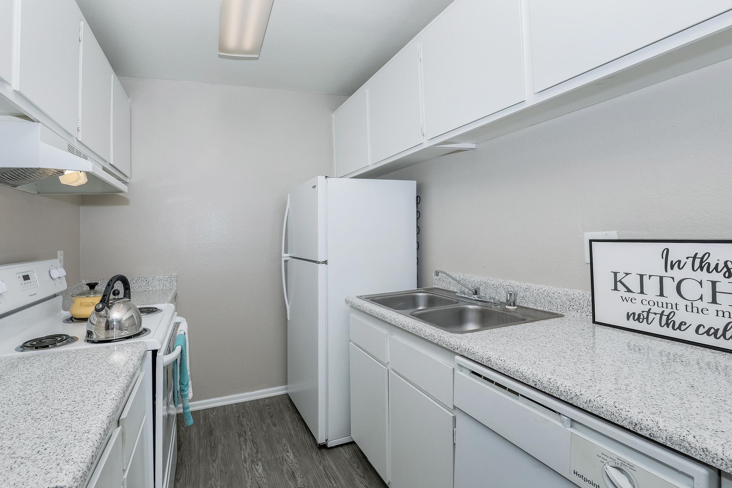 Two Bedroom Apartments in San Bernardino CA - Creekside Village - Kitchen with Wood-Style Vinyl Flooring, White Cabinetry, Double Sink, White Appliances, and Overhead Lighting.