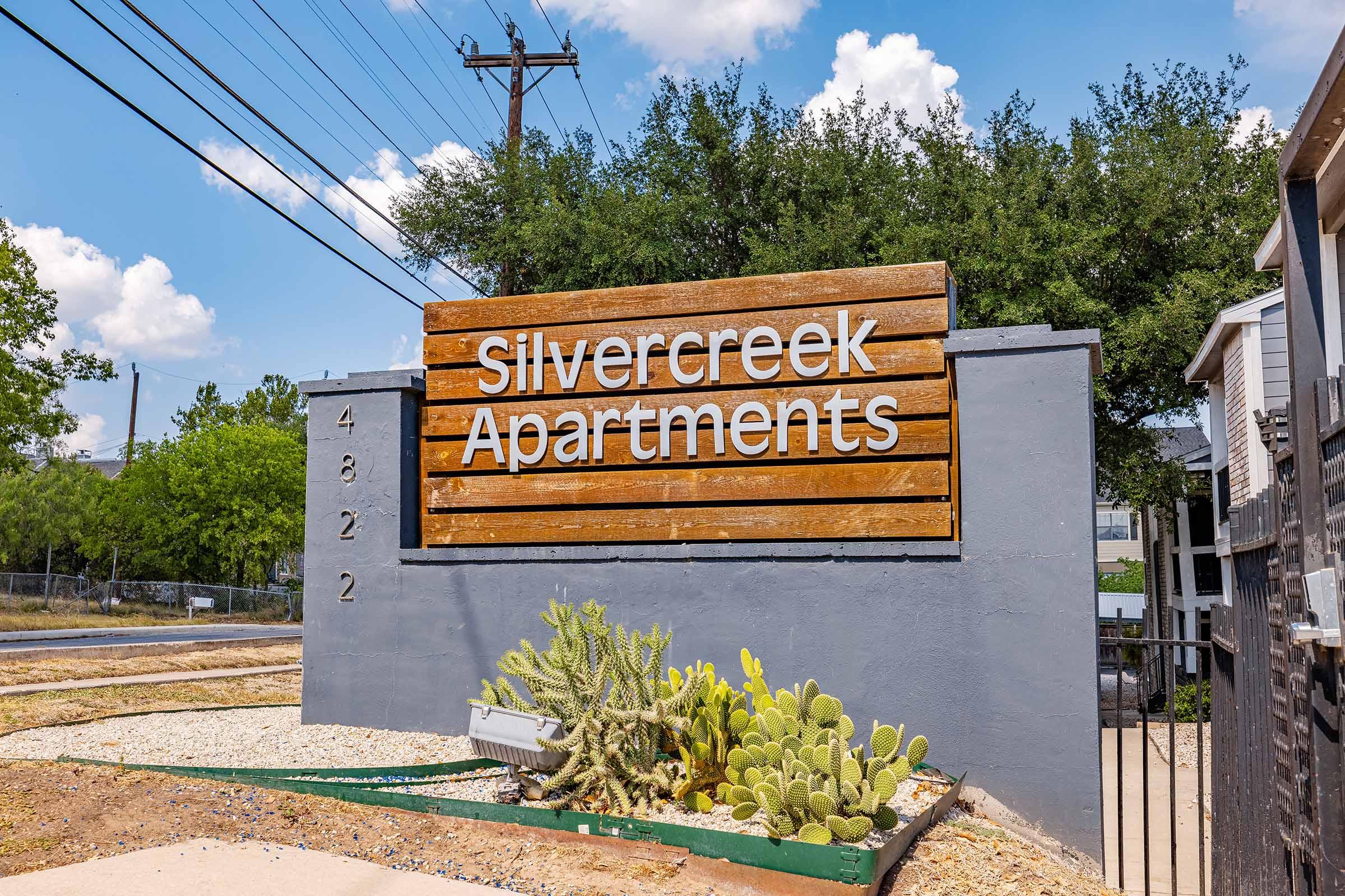 Sign for Silvercreek Apartments with a wooden design, featuring the name prominently displayed. Surrounding greenery includes cacti and other plants, set against a clear blue sky with a few clouds. Electrical poles are visible in the background.