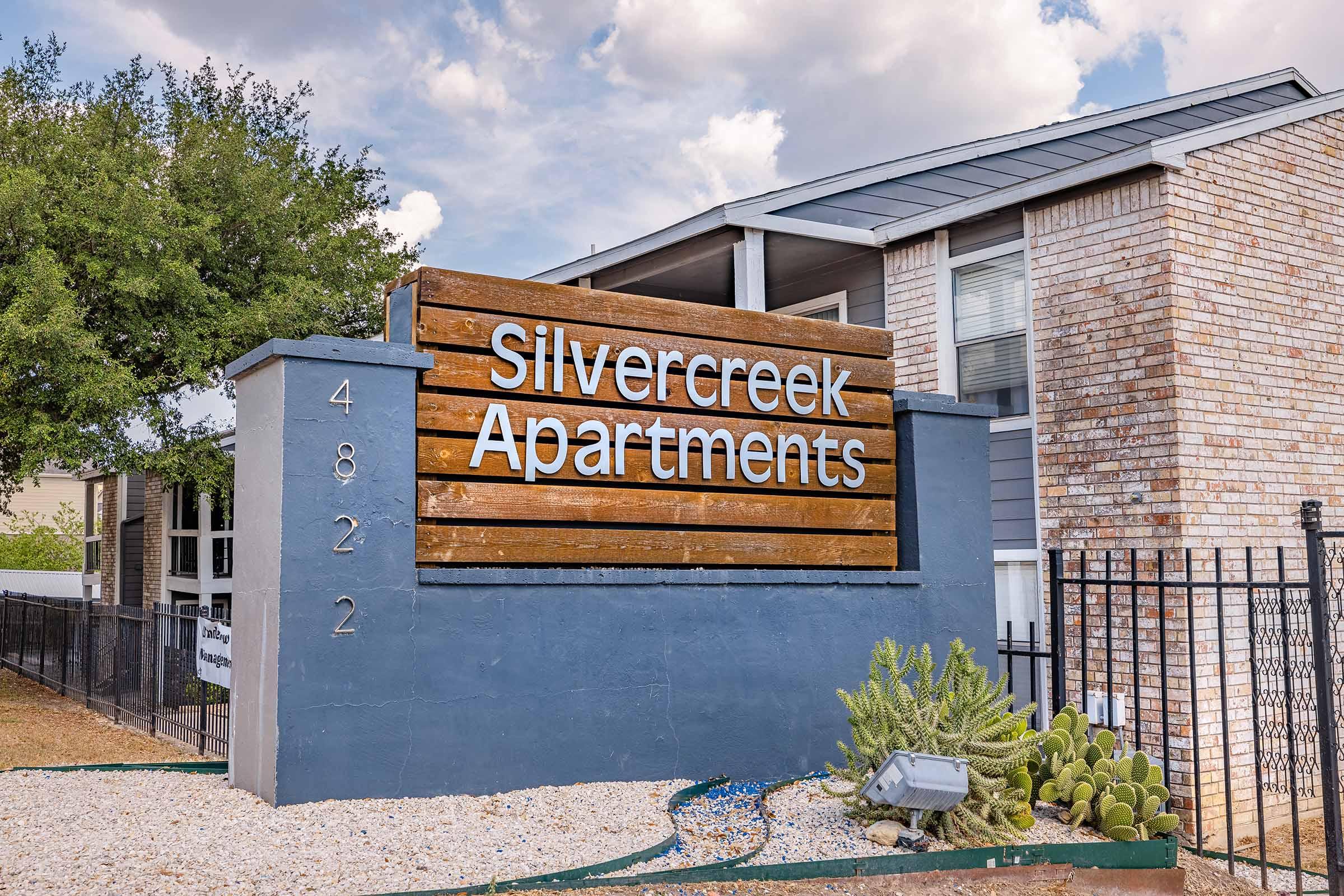 Sign for Silvercreek Apartments at 4822, featuring wooden panels with white lettering against a backdrop of a modern apartment building. Surrounding area includes gravel and desert plants, with a clear sky and trees in the background.