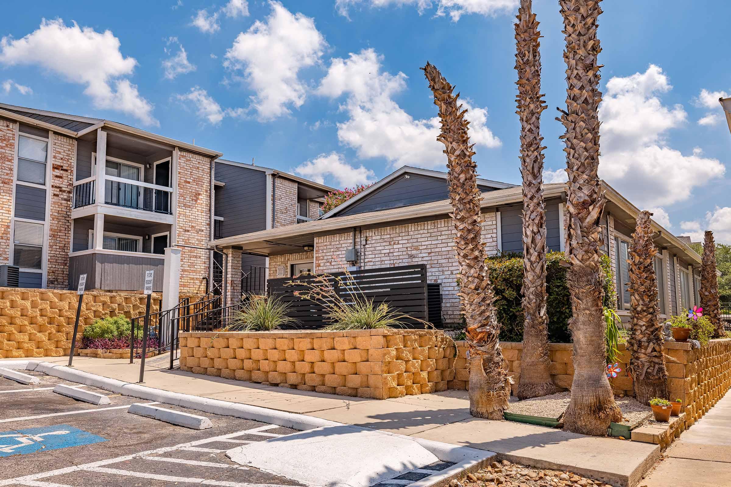 A modern apartment building with a landscaped front area featuring palm trees and decorative plants. The building has a mix of brick and wood siding, with balconies on the upper floor. In the foreground, there is a parking area marked with lines and a wheelchair-accessible space. Bright blue sky with fluffy clouds above.