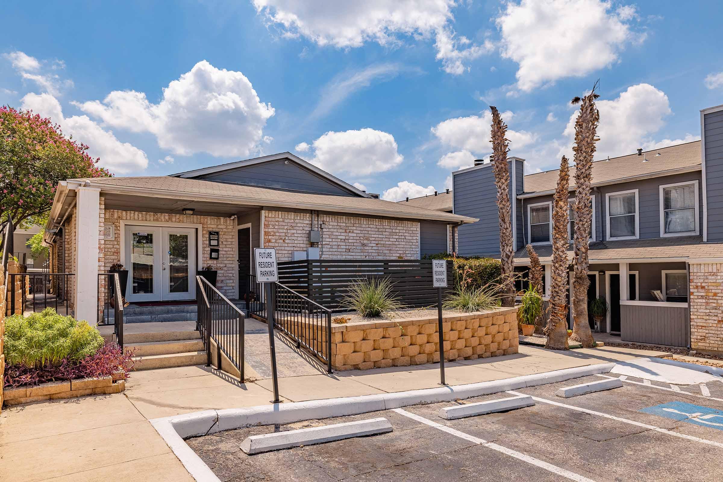 A well-maintained apartment building with a brick facade, featuring a welcoming entrance and landscaped area. Palm trees are visible, alongside a parking lot with clearly marked spaces. The sky is bright blue with fluffy white clouds, creating a sunny atmosphere.