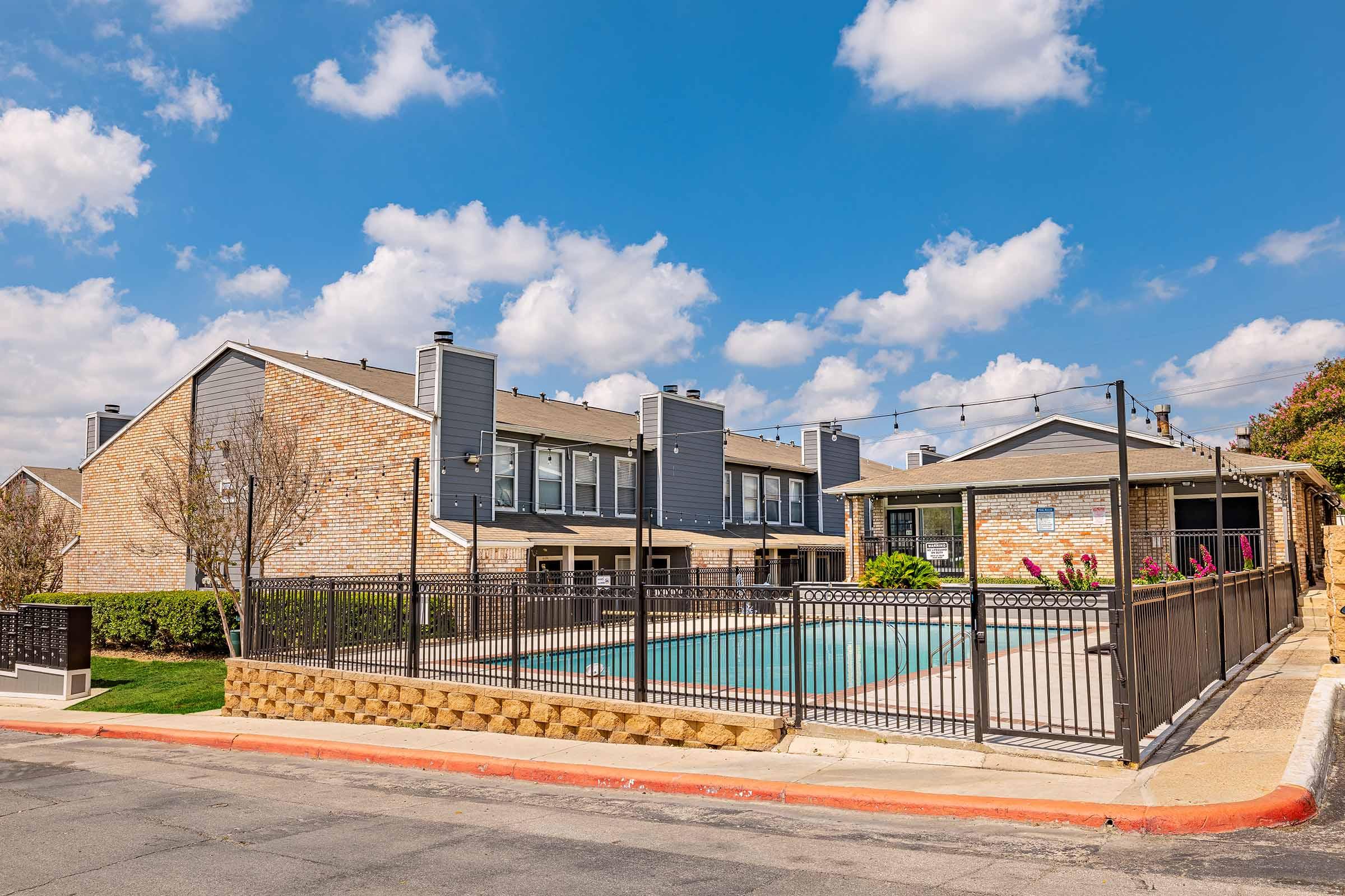 A residential area featuring a swimming pool surrounded by a black fence. The buildings nearby have brick exteriors and large windows. Blue skies with fluffy white clouds are visible above, creating a bright and inviting atmosphere.